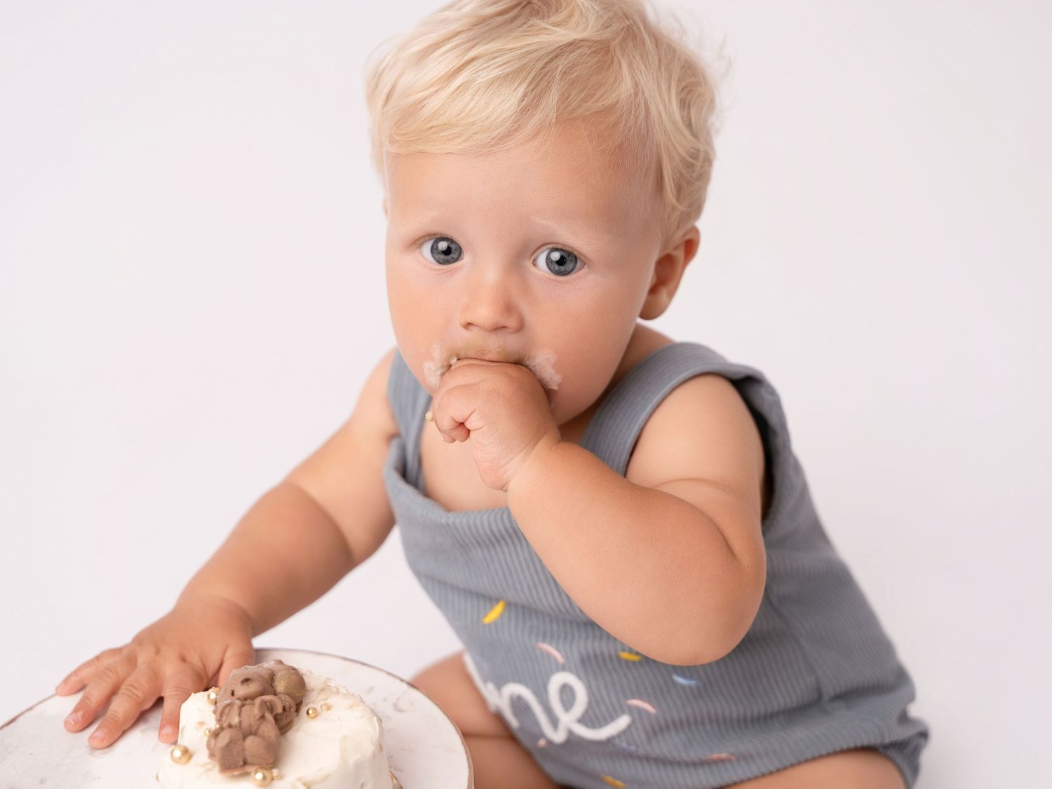 Blonde toddler in gray romper eating cake with frosting on face during first birthday smash cake photo session.