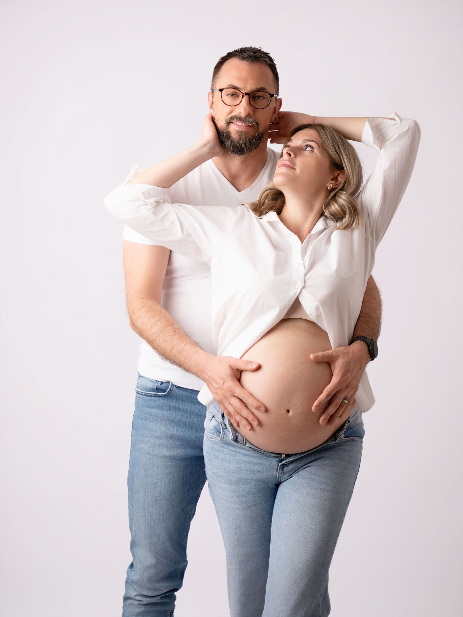 Couple in matching white shirts and jeans posing together, pregnant woman cradling baby bump while partner embraces her from behind.