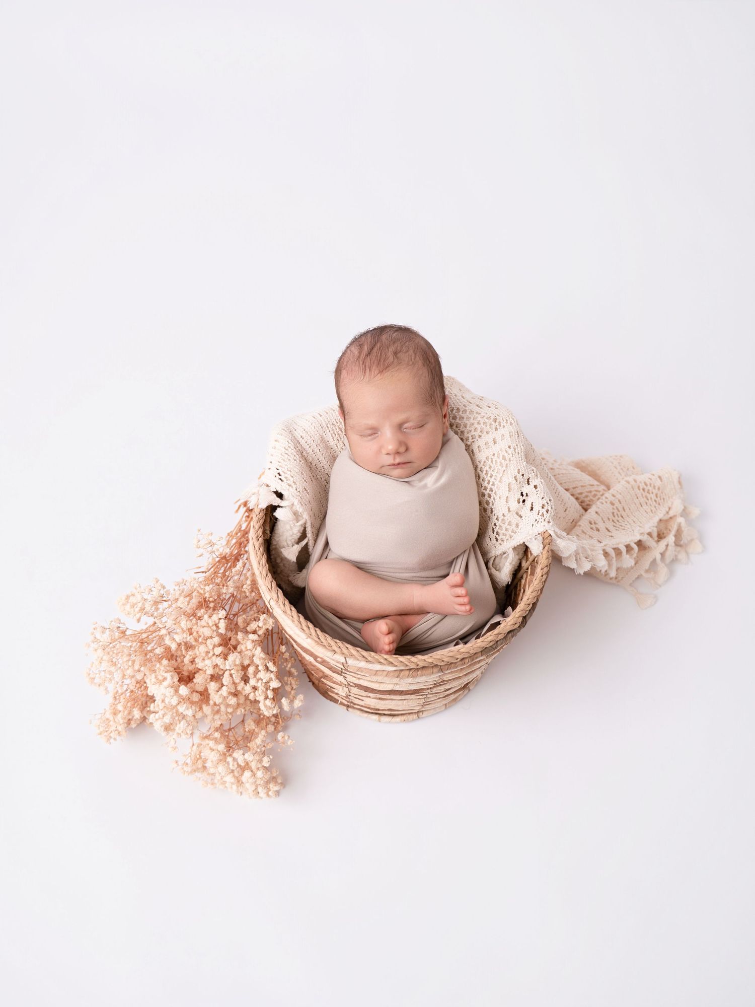 Newborn baby sleeping peacefully in a wicker basket with cream knit blanket and soft pink dried flowers.