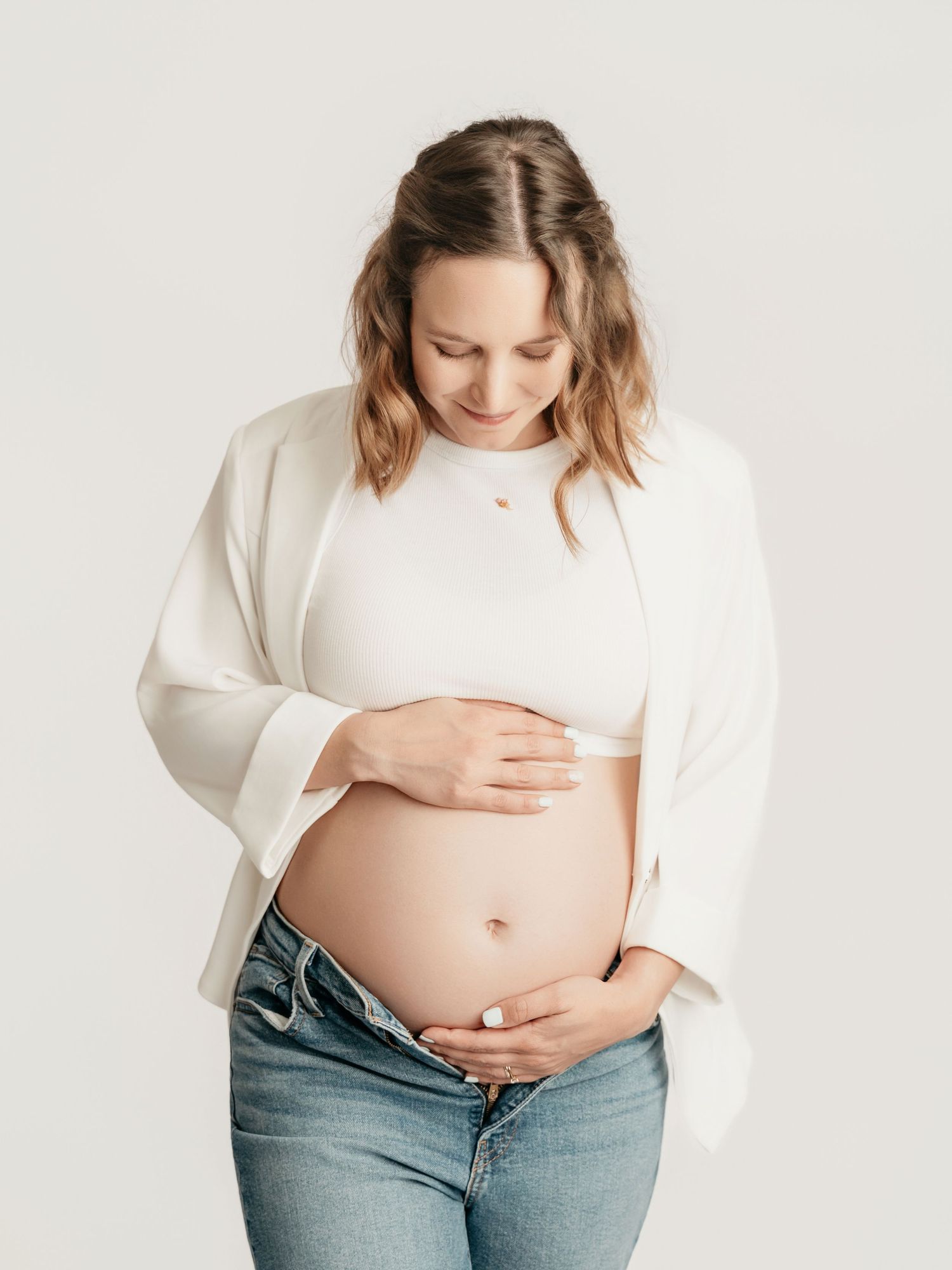 Smiling pregnant woman in white crop top and blazer gently holding bare baby bump in studio.