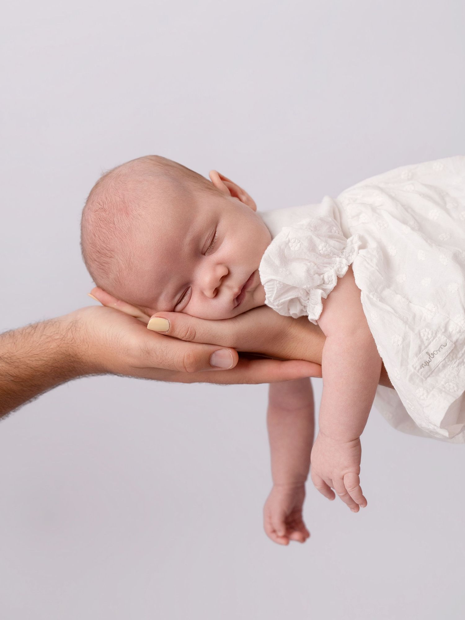 Newborn baby sleeping peacefully on father's hand, dressed in white, against a soft gray background.