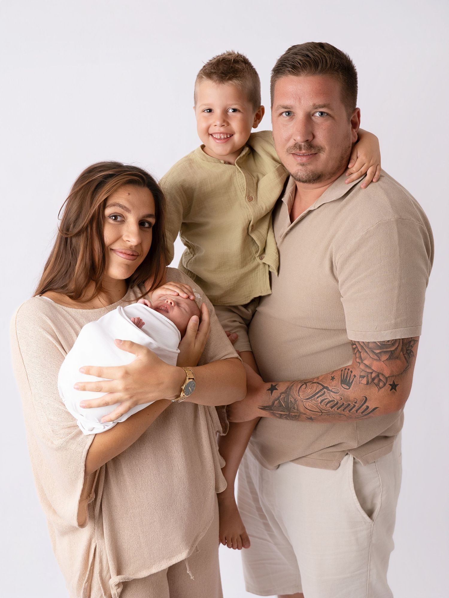 Family studio portrait of parents with newborn baby and young son, dressed in coordinating neutral beige outfits.