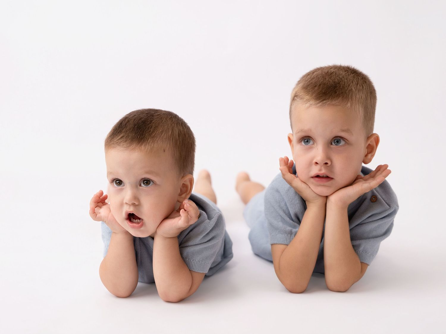 Two young brothers in blue outfits lying on white background, resting chins on hands, looking at camera.