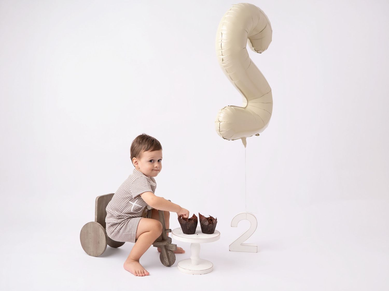 Toddler boy sitting near chocolate cupcakes with a number 2 balloon, celebrating his 2nd birthday.