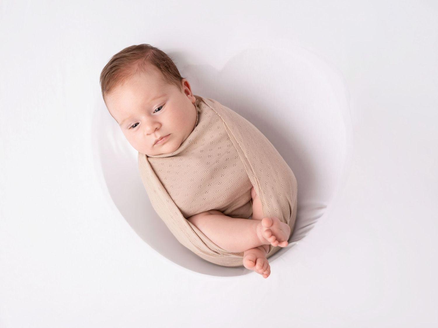 Newborn baby swaddled in beige wrap sleeping peacefully in a white bowl on a white background.