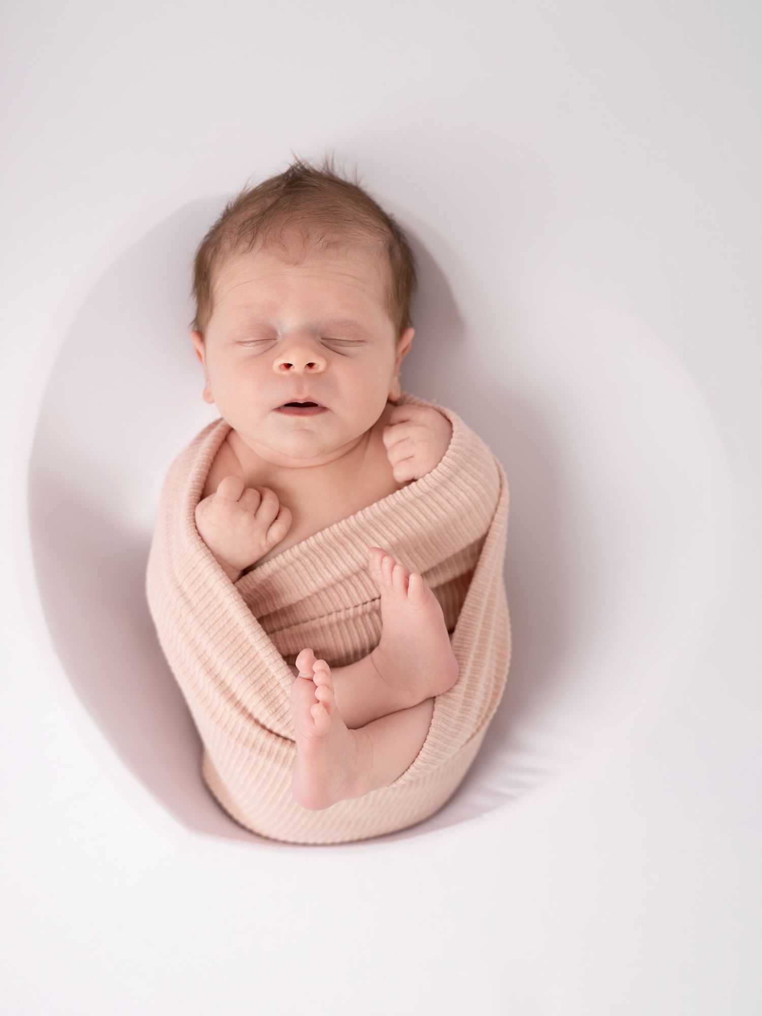 Peaceful newborn baby swaddled in a soft pink knit wrap, sleeping in a round white bowl prop.
