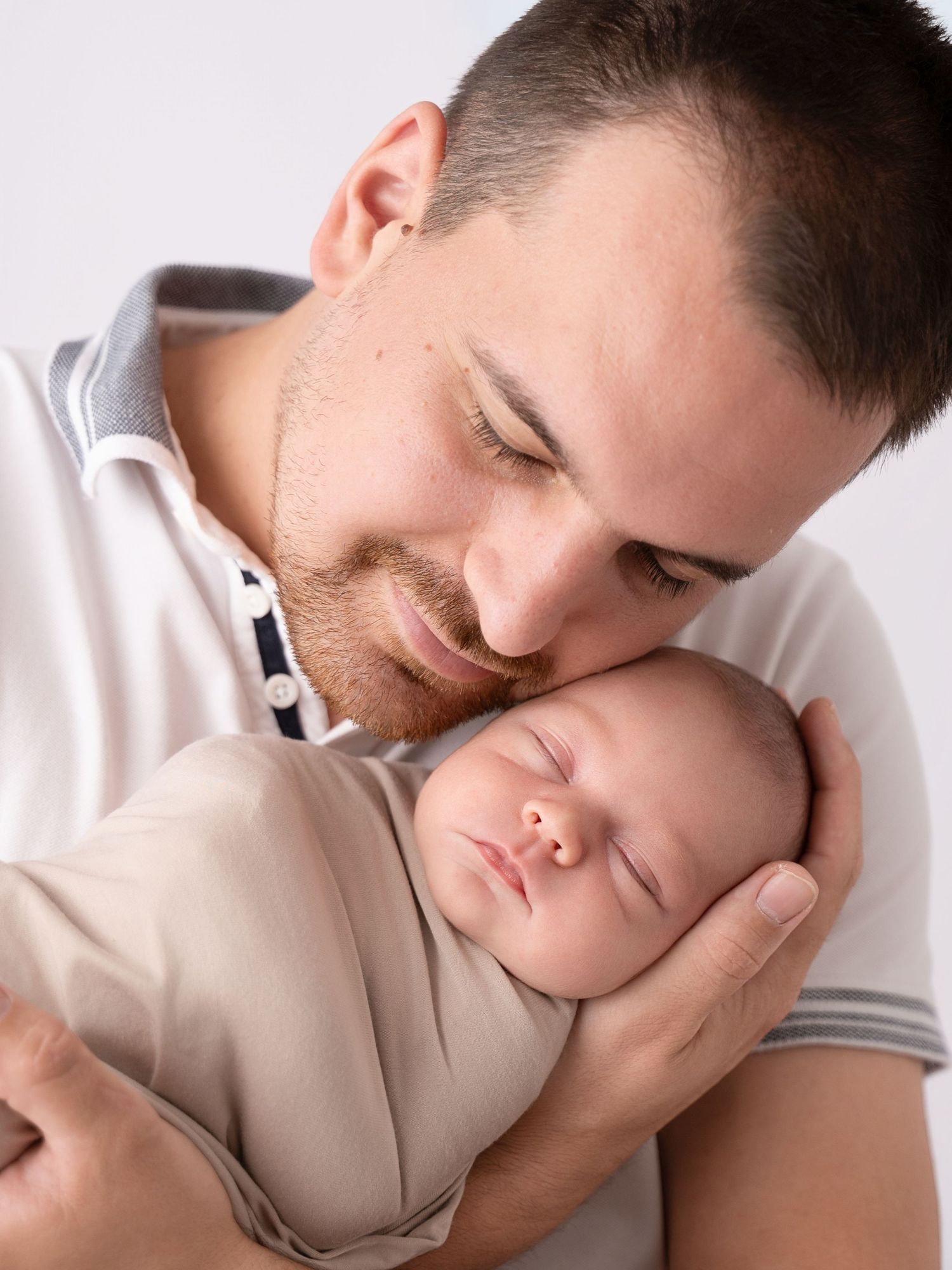 A loving father gently holds and nuzzles his sleeping newborn baby wrapped in a soft beige swaddle.