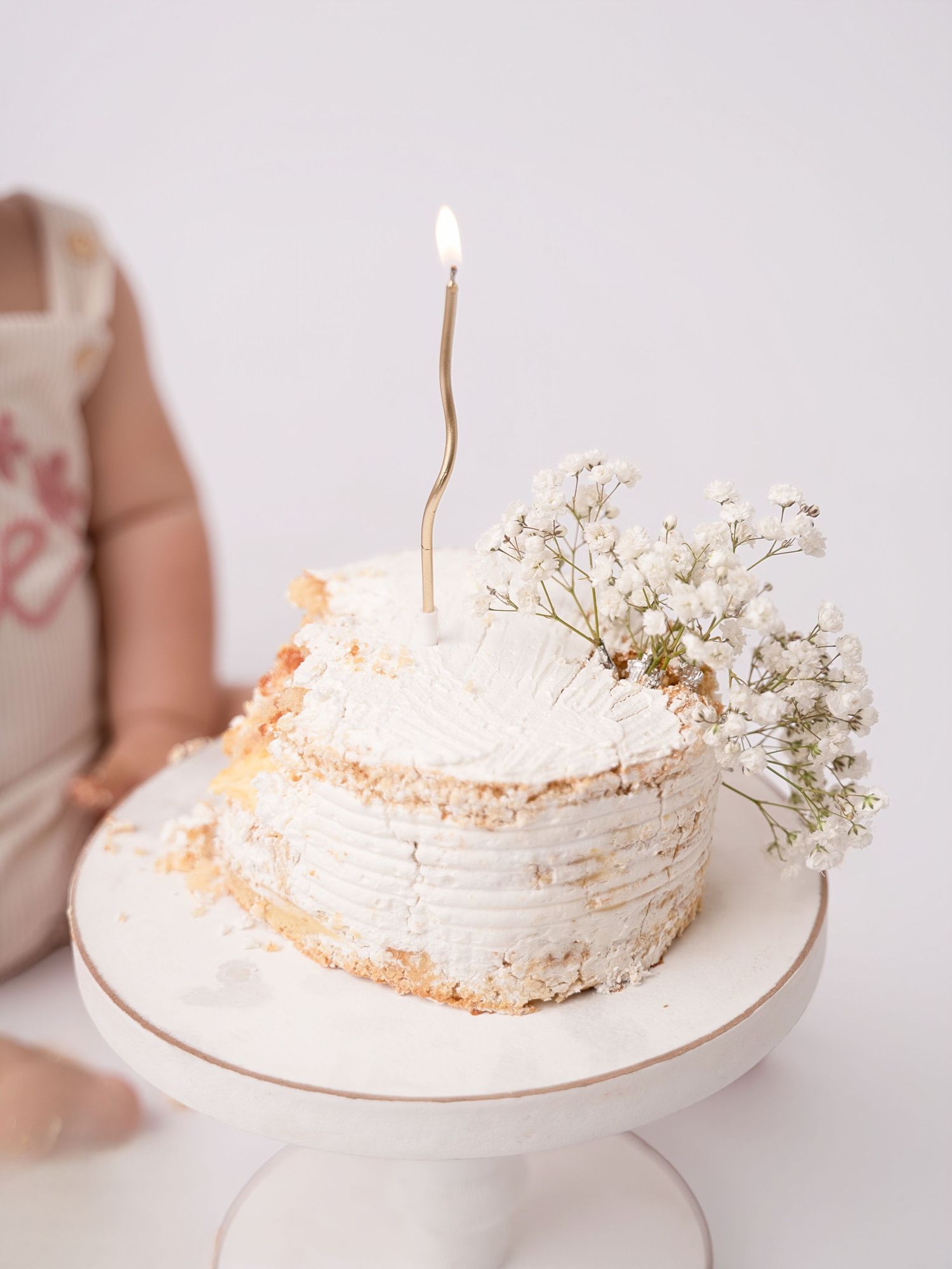 White frosted birthday smash cake with a single gold candle and baby's breath flowers on a white cake stand.