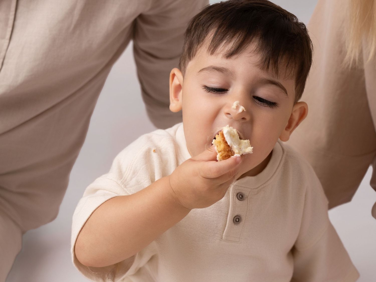 Toddler boy in cream outfit eating a snack, held by parent in bright studio setting.
