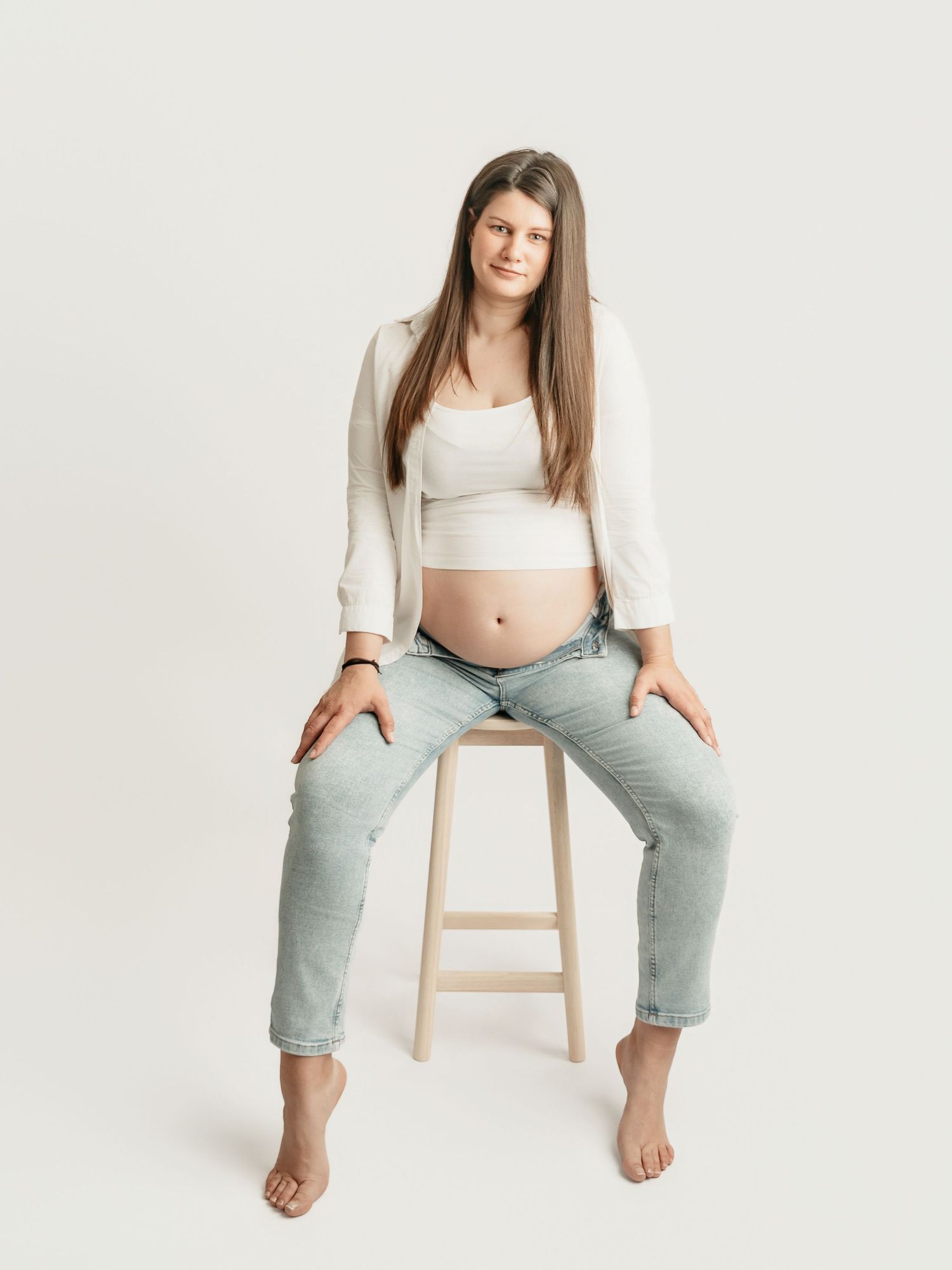 Pregnant woman in white crop top and jeans sitting on stool showing baby bump in studio portrait.