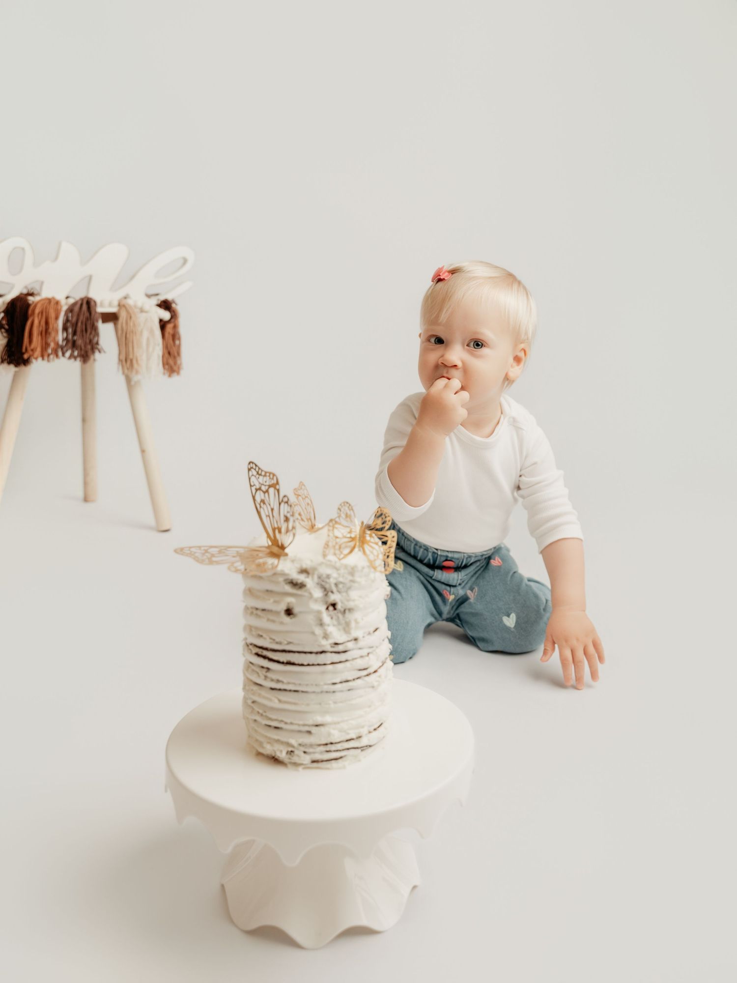 An adorable blonde toddler sits beside a white layered birthday cake in a bright studio.