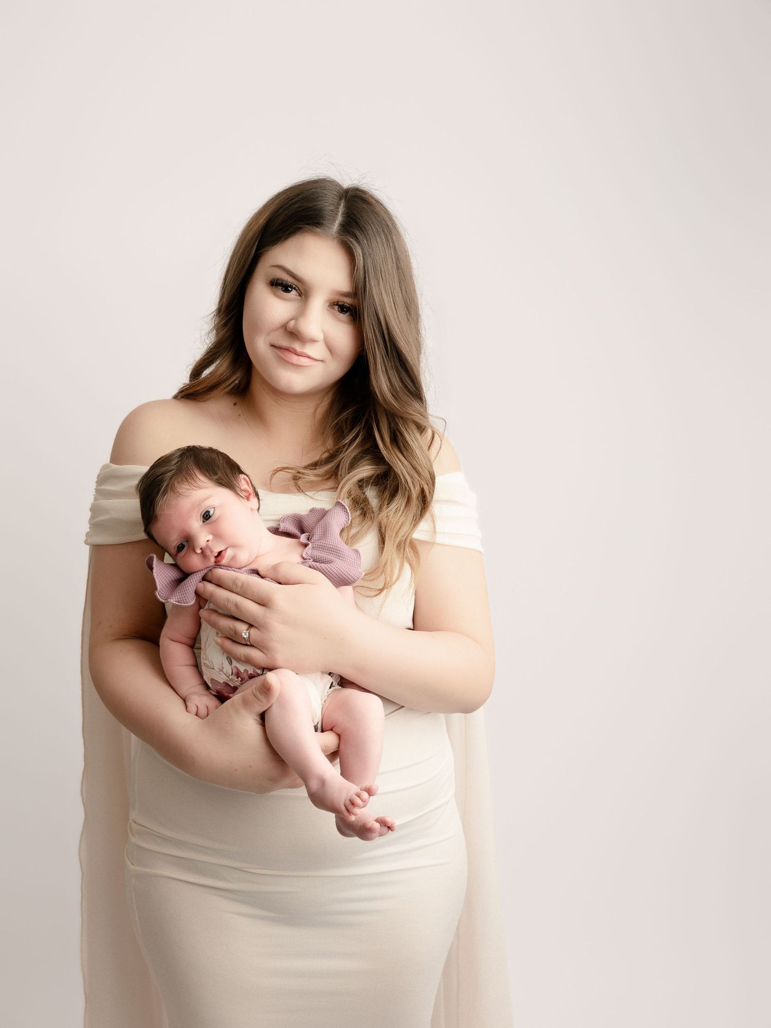 Young mother in cream off-shoulder gown holds newborn baby in pink outfit against white studio background.