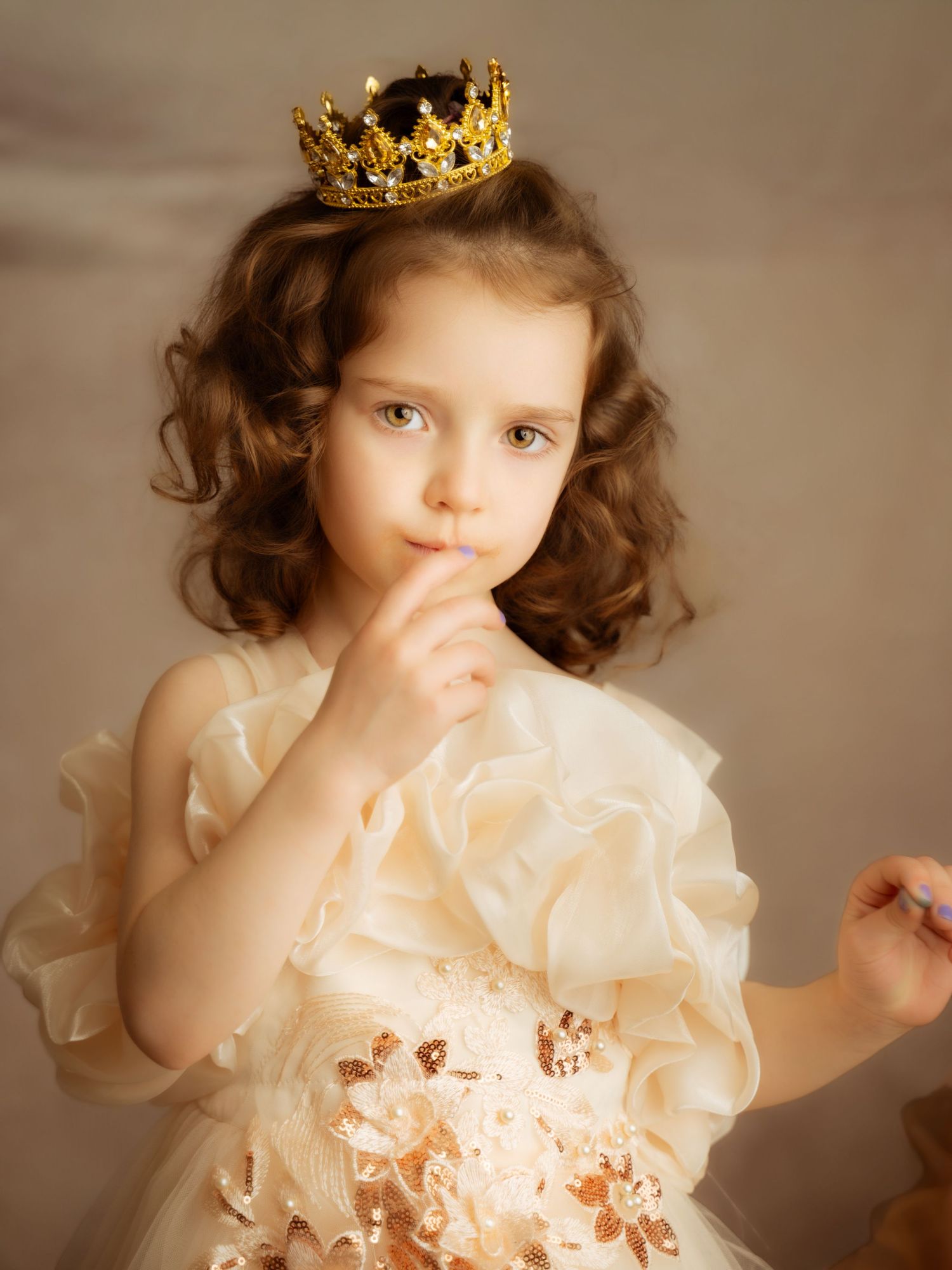 Curly-haired little girl wearing gold crown in cream ruffled dress with thoughtful expression.
