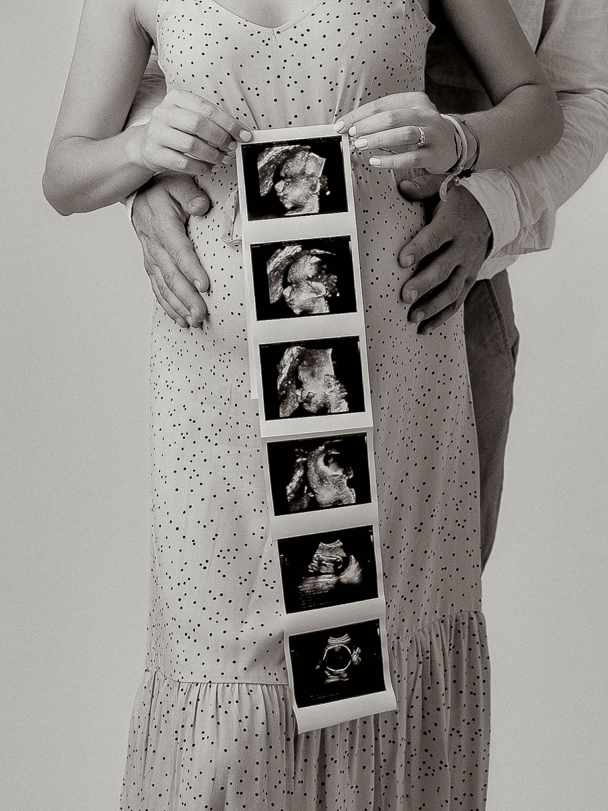 Black and white photo of pregnant couple holding ultrasound strip showing baby scan images, maternity announcement.