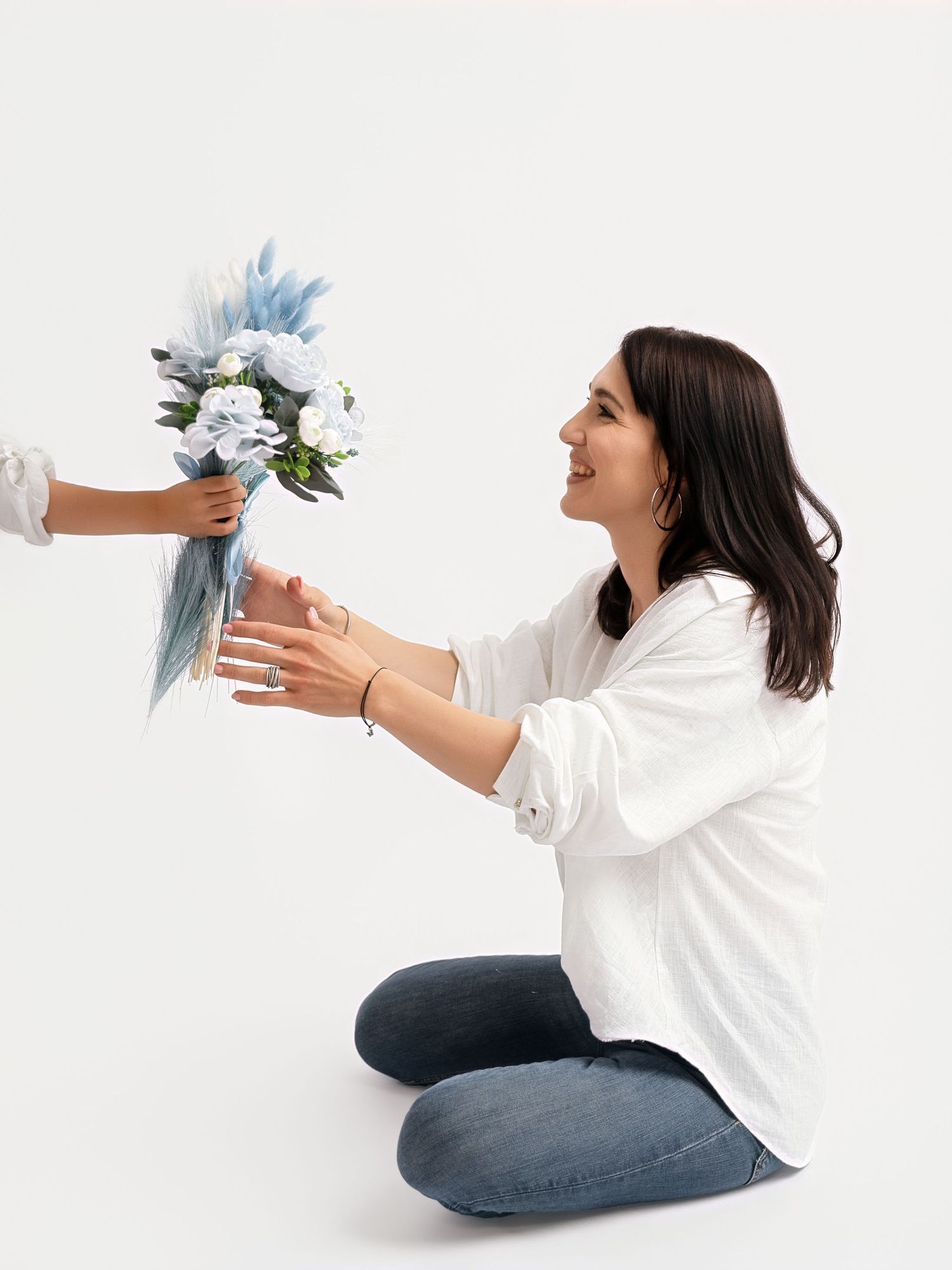 A smiling woman in a white shirt receives a bouquet of blue and white flowers while sitting on the floor.
