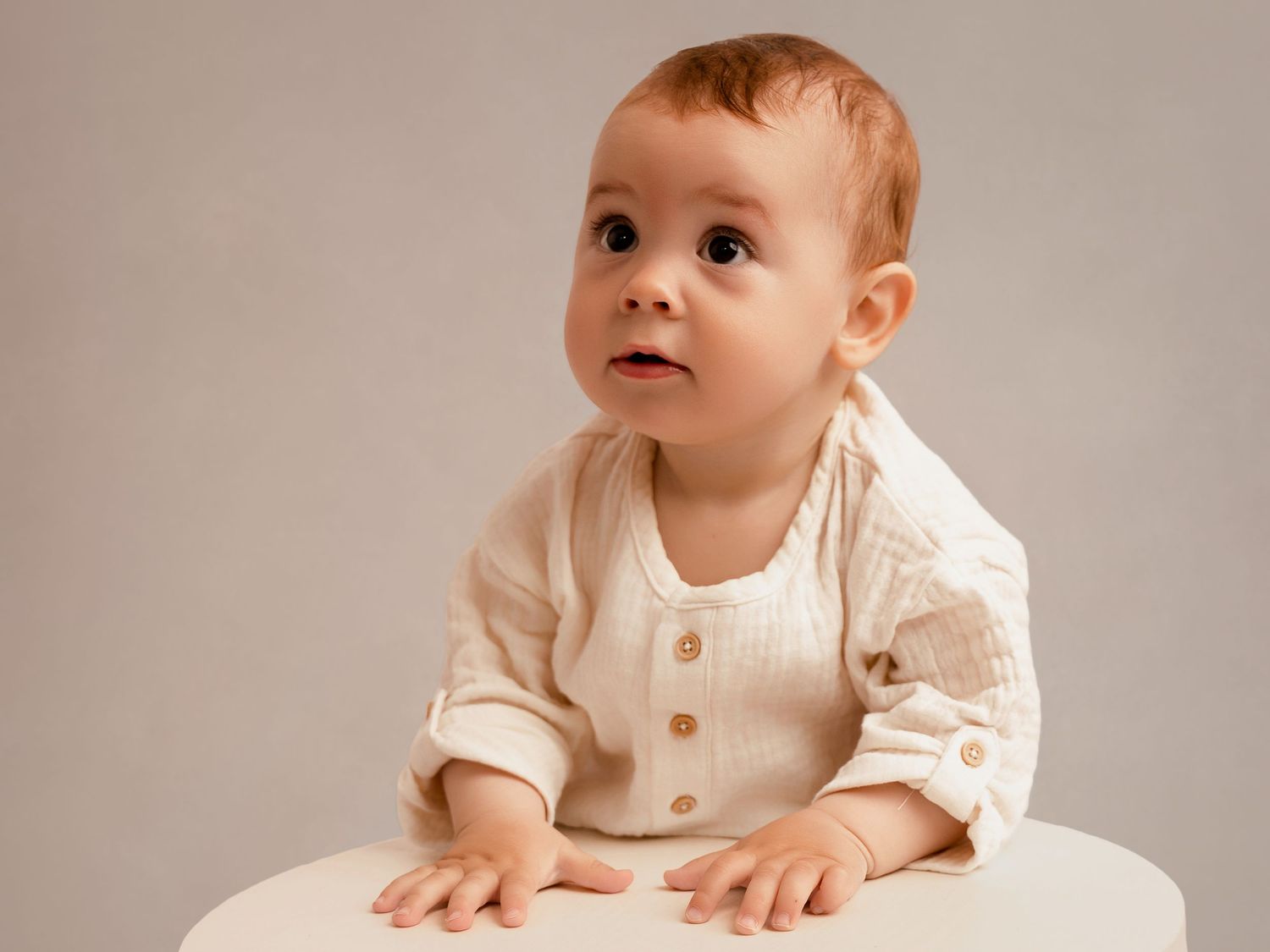 A cute baby in a cream linen outfit sits at a white table, looking up curiously against a neutral background.