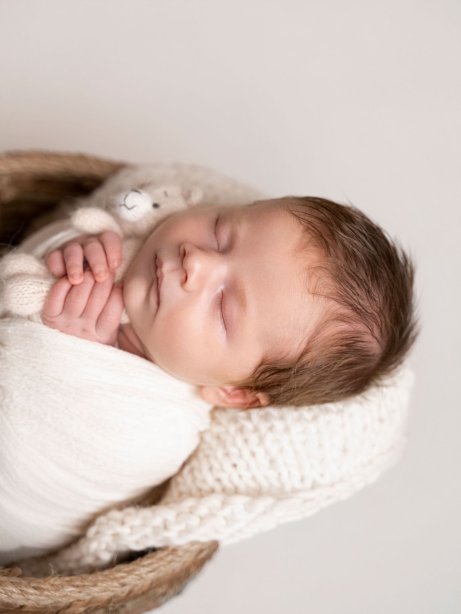 Peaceful newborn baby sleeping in a woven basket, wrapped in a cream knit blanket, holding a tiny stuffed animal.