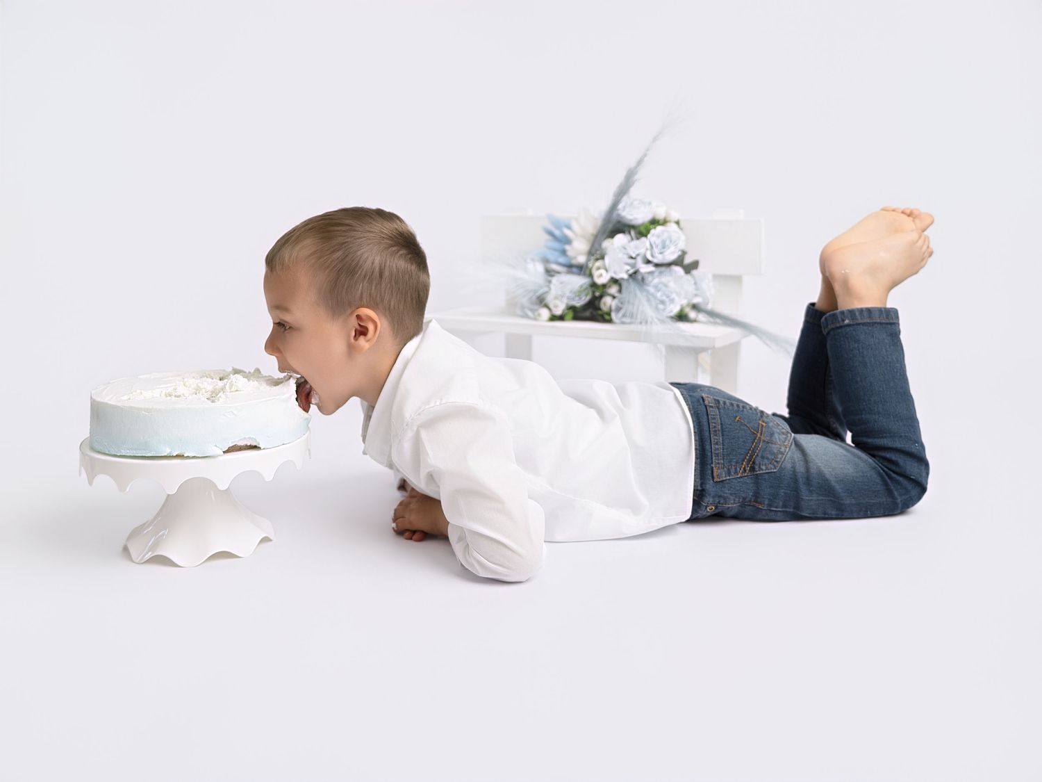 Young boy in white shirt and jeans lying on floor eagerly eating a small white birthday cake.