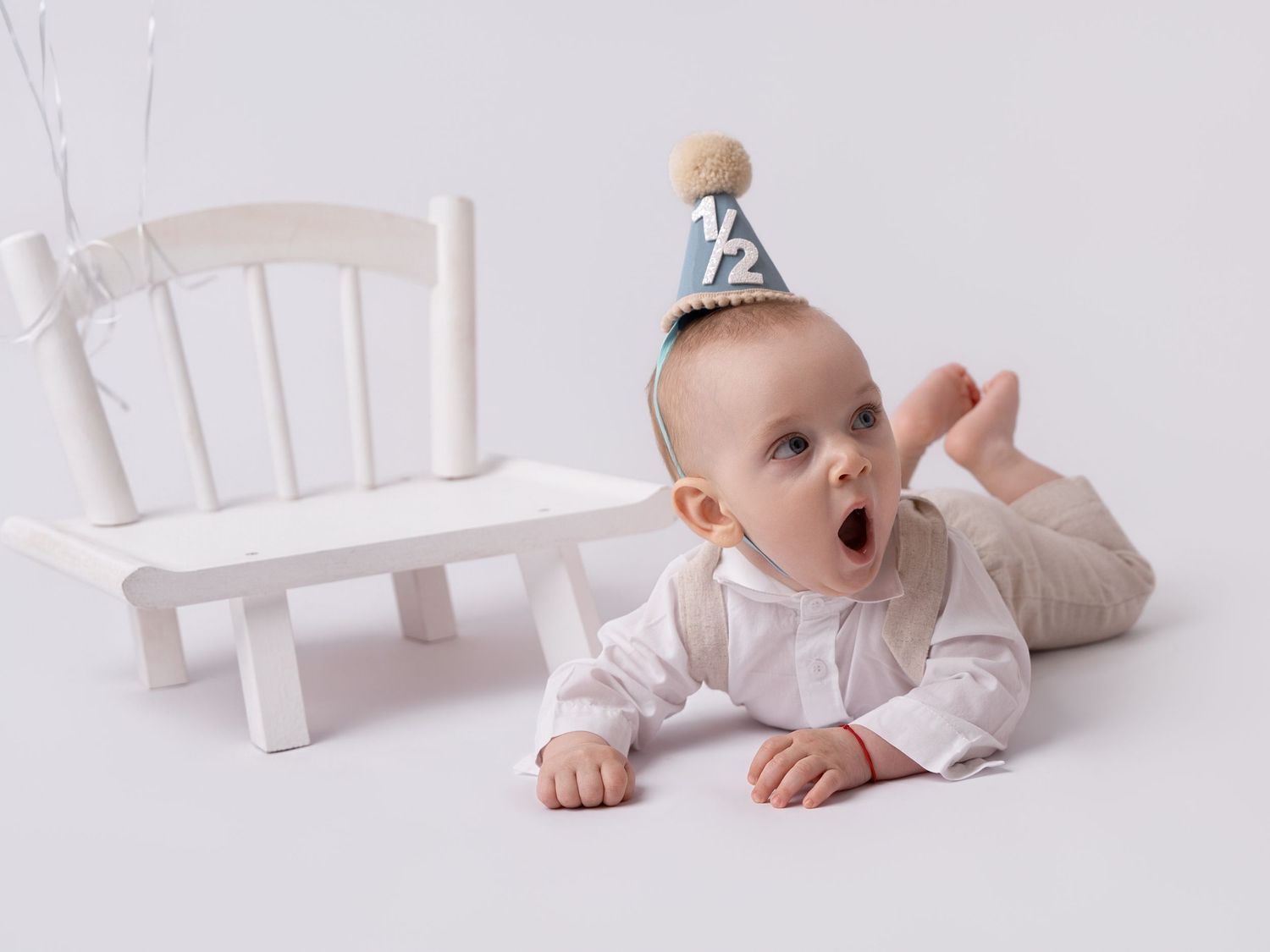 Adorable baby wearing a half birthday party hat lying on white floor next to a small white bench.