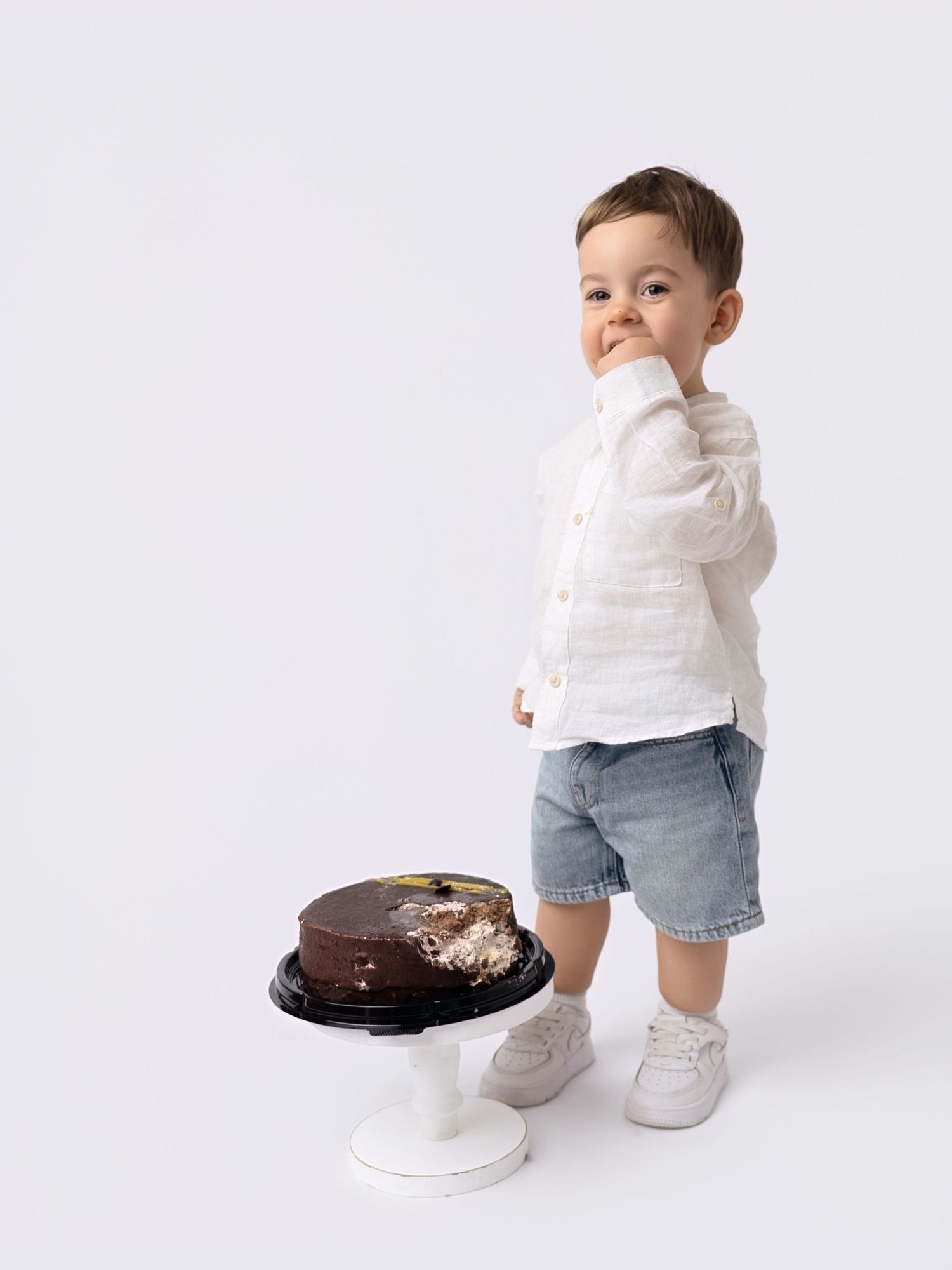 Cute toddler boy in white linen shirt and denim shorts standing next to a chocolate smash cake.