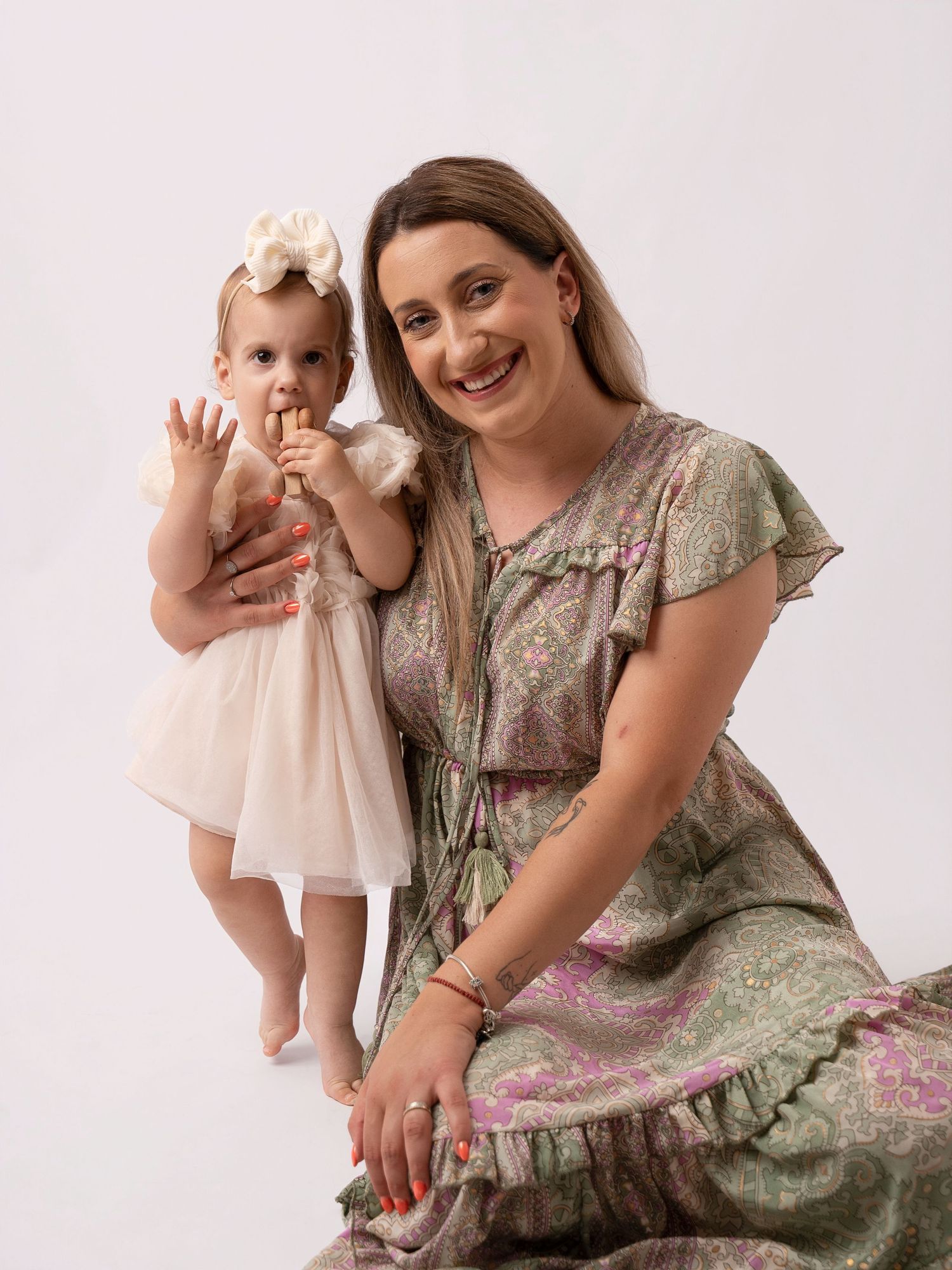 A smiling woman in a green floral dress sits with a baby girl in a white tutu dress holding a treat.