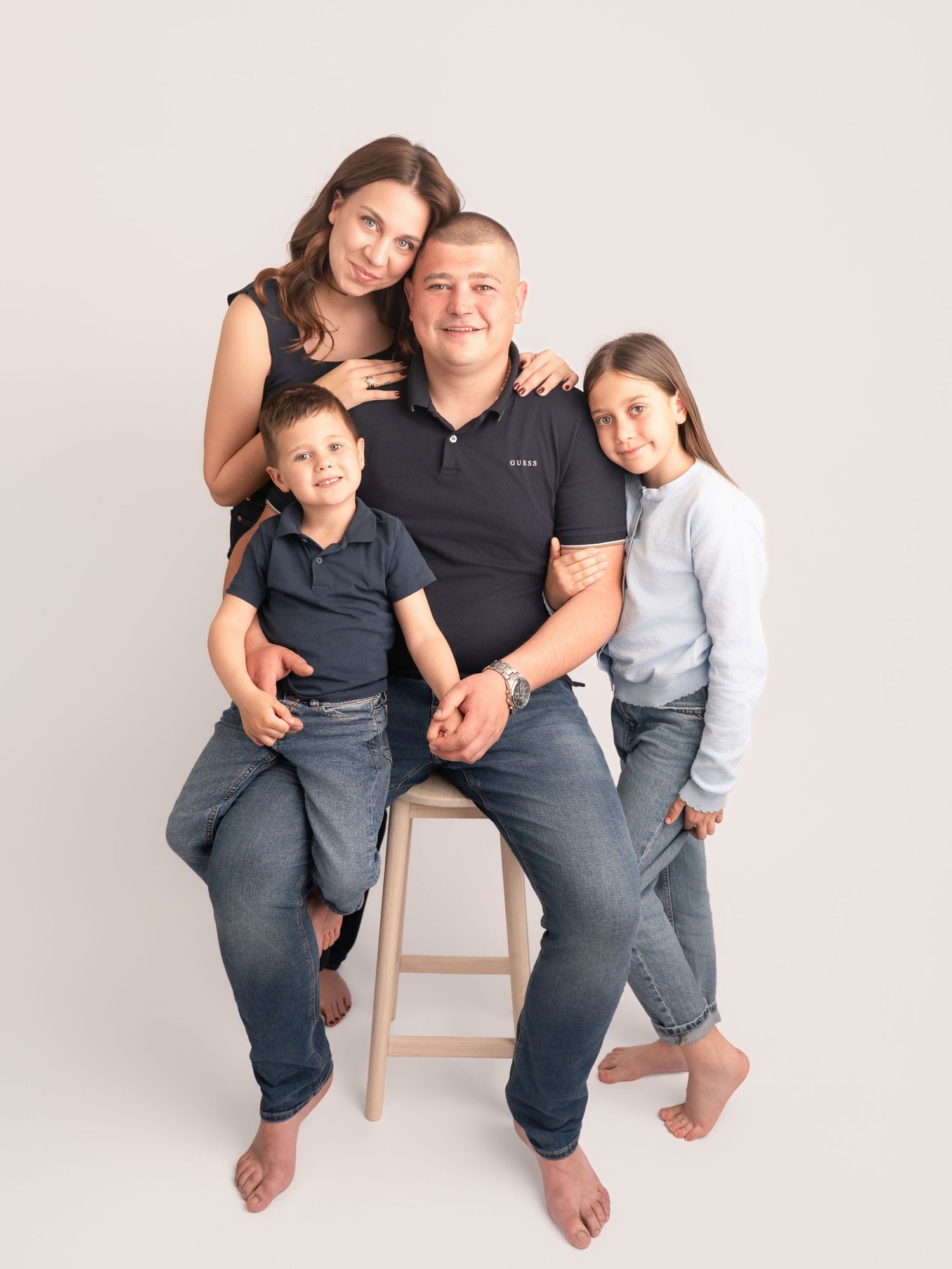 Happy family of four posing together on a stool in a bright professional photo studio.