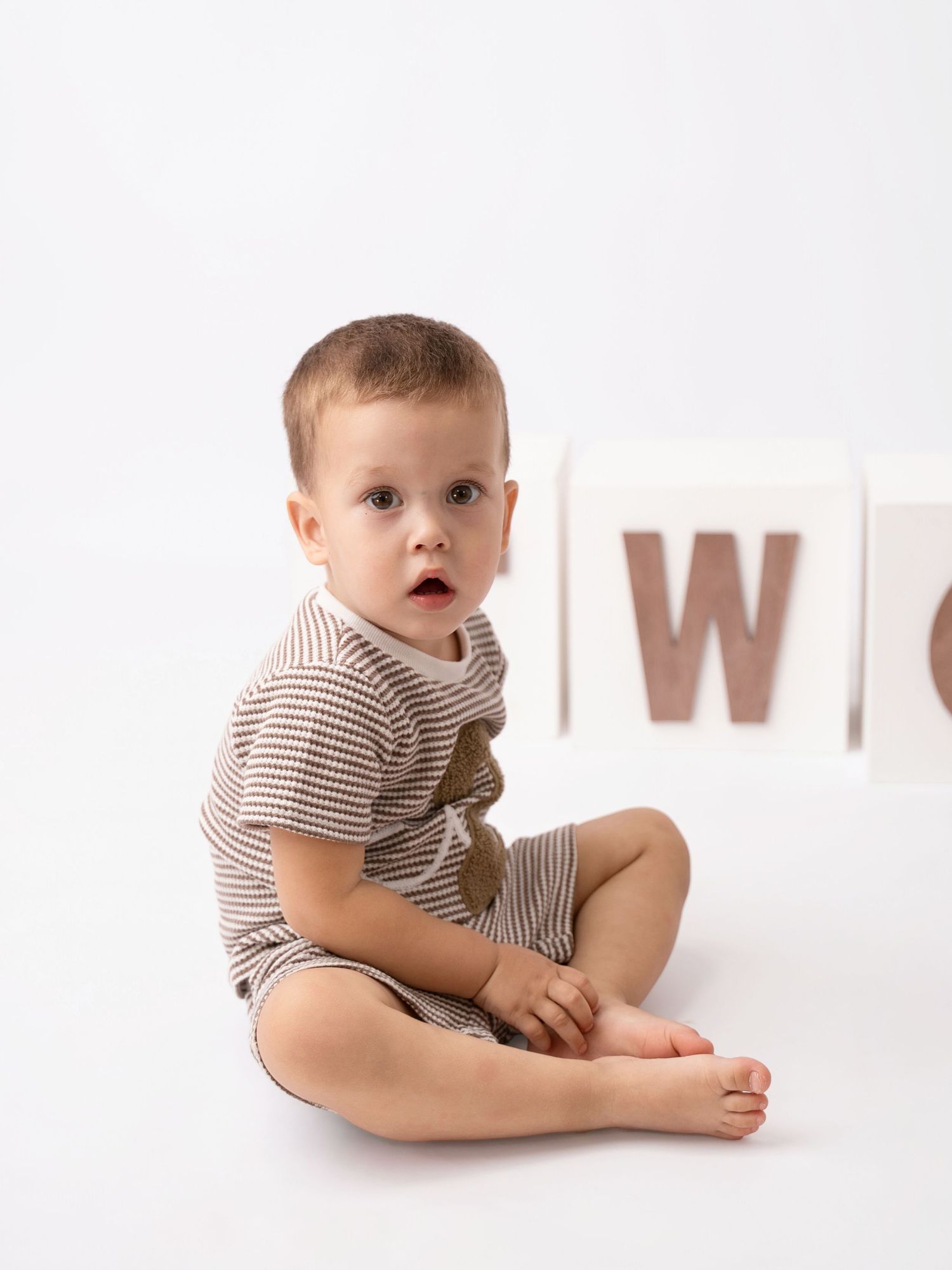 Cute baby boy in striped brown romper sitting on white floor next to wooden alphabet blocks.