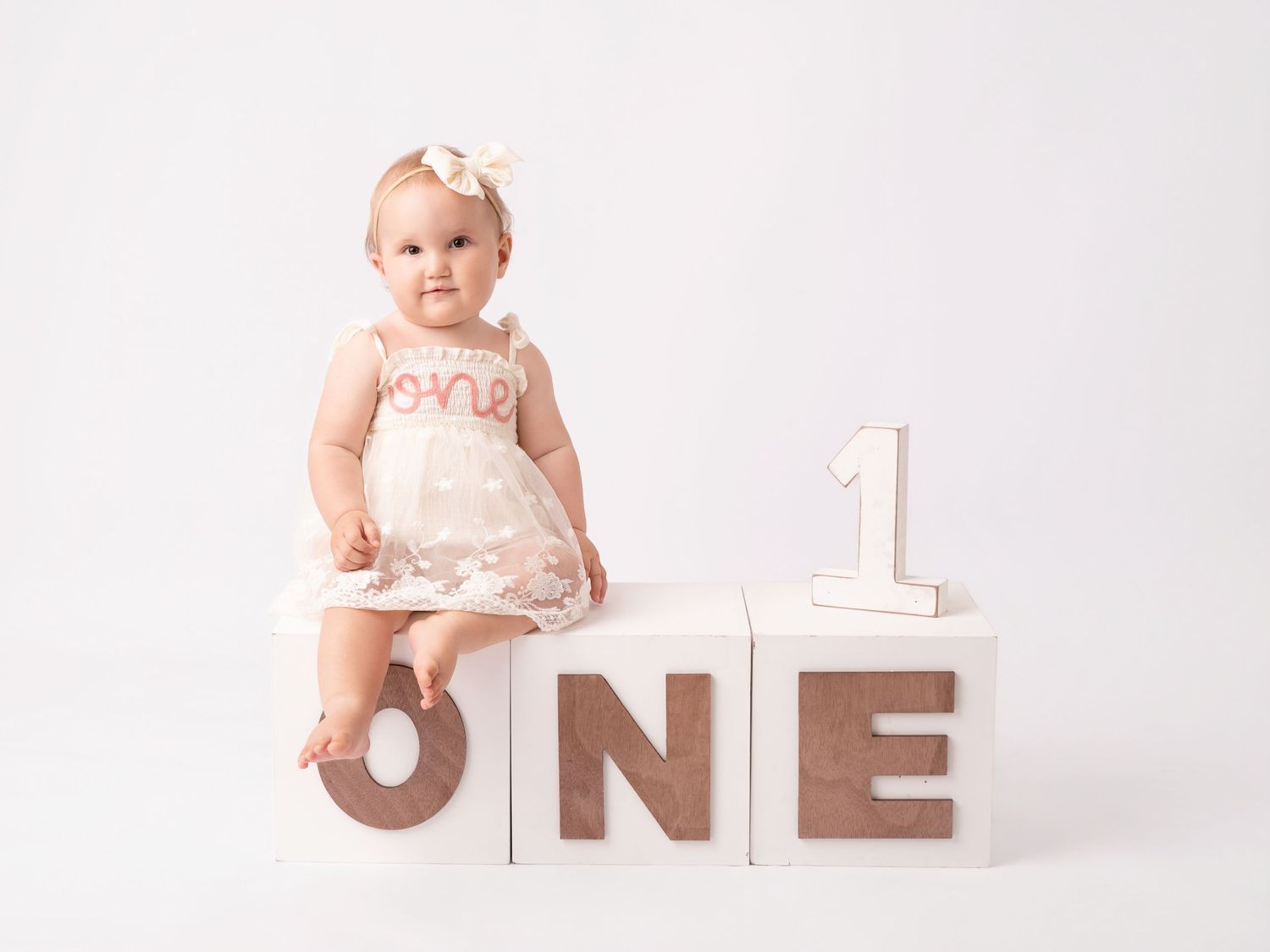 Baby girl in white dress sits on ONE blocks celebrating her first birthday in a bright studio photo.