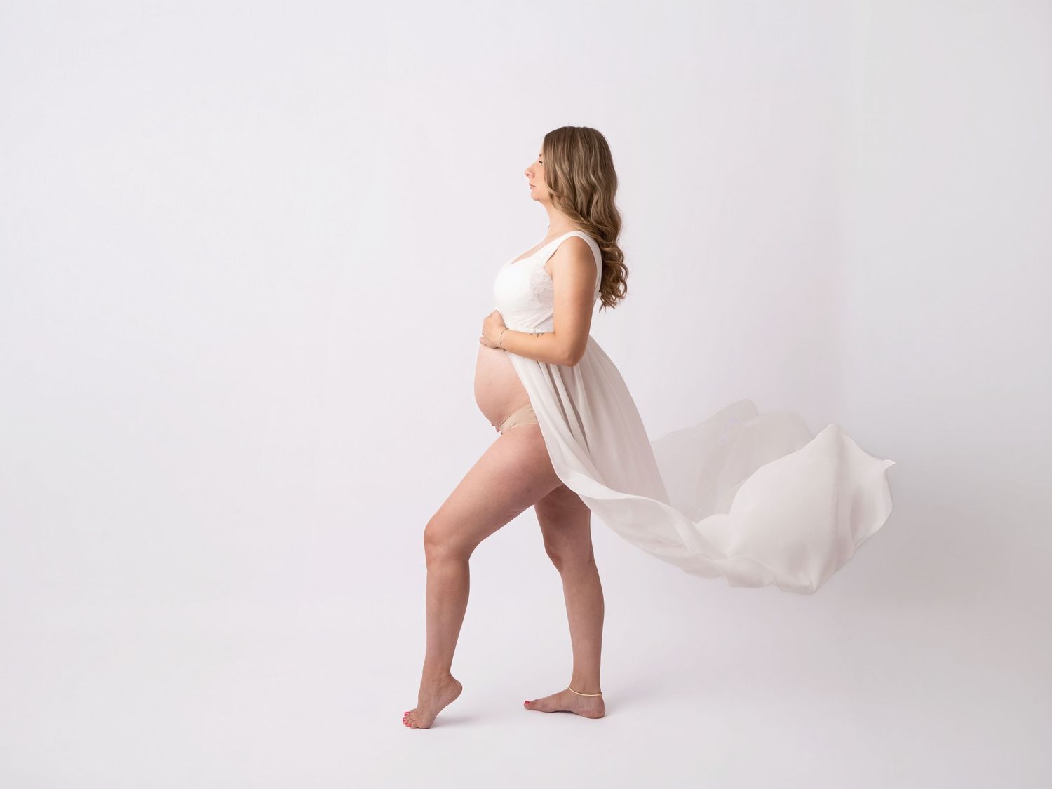Pregnant woman in white top with flowing fabric against white background, cradling baby bump in elegant maternity photo.