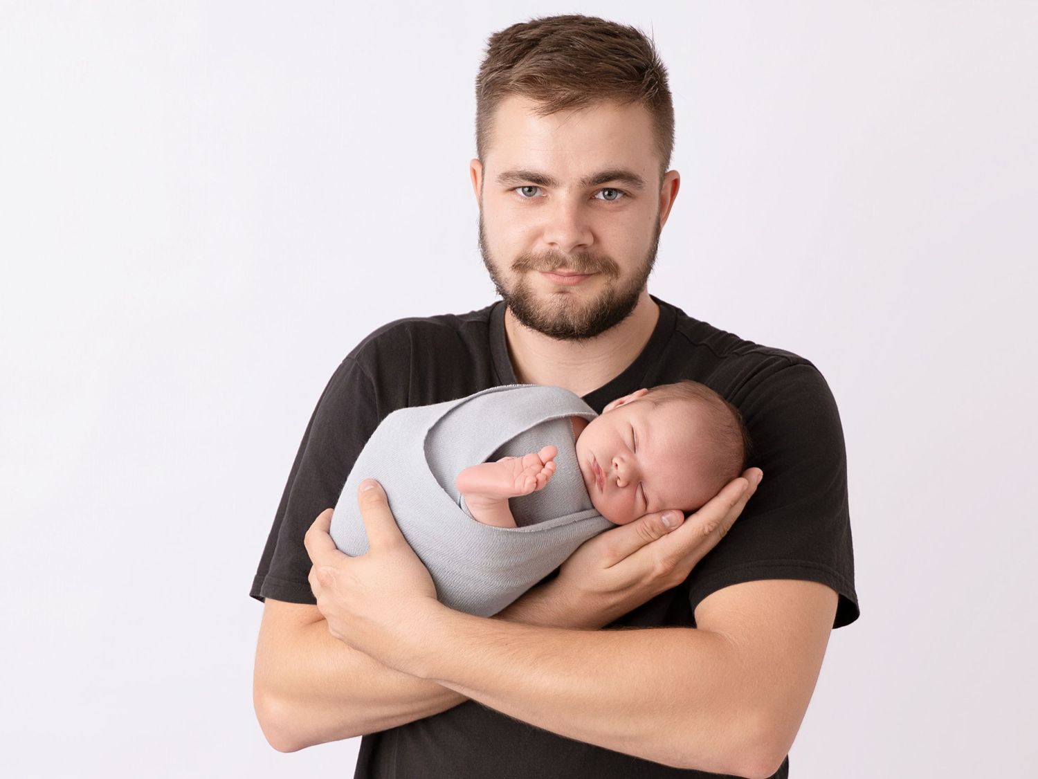 Young bearded man in black t-shirt tenderly holding a sleeping newborn baby wrapped in gray cloth.