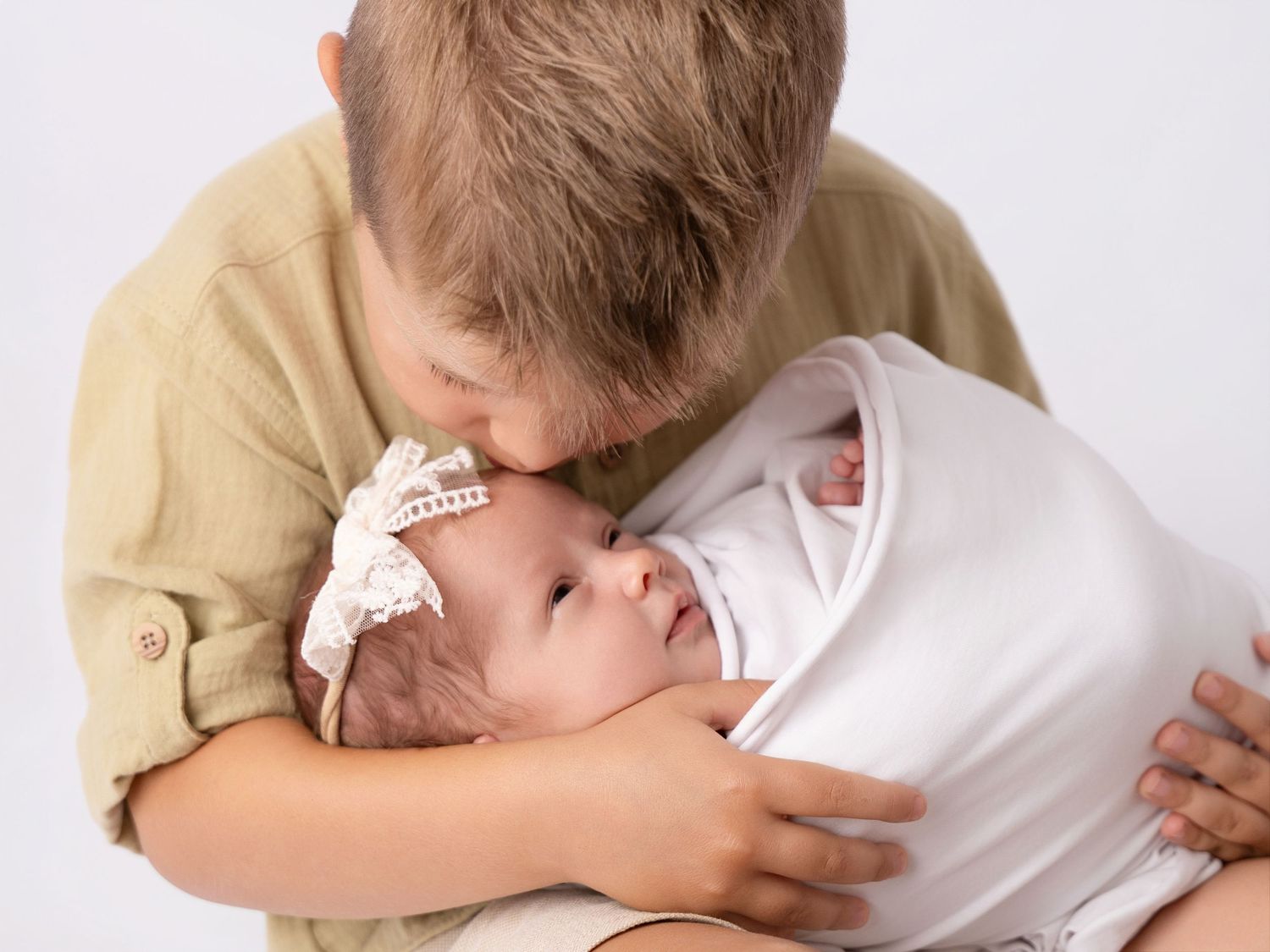 Older boy gently kissing newborn baby sister wrapped in white, wearing lace headband, in a sweet sibling portrait.