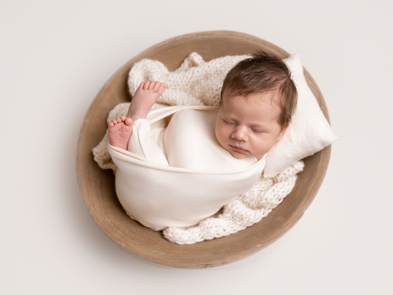 Newborn baby sleeping peacefully in a wooden bowl, wrapped in a cream swaddle with a knit blanket.
