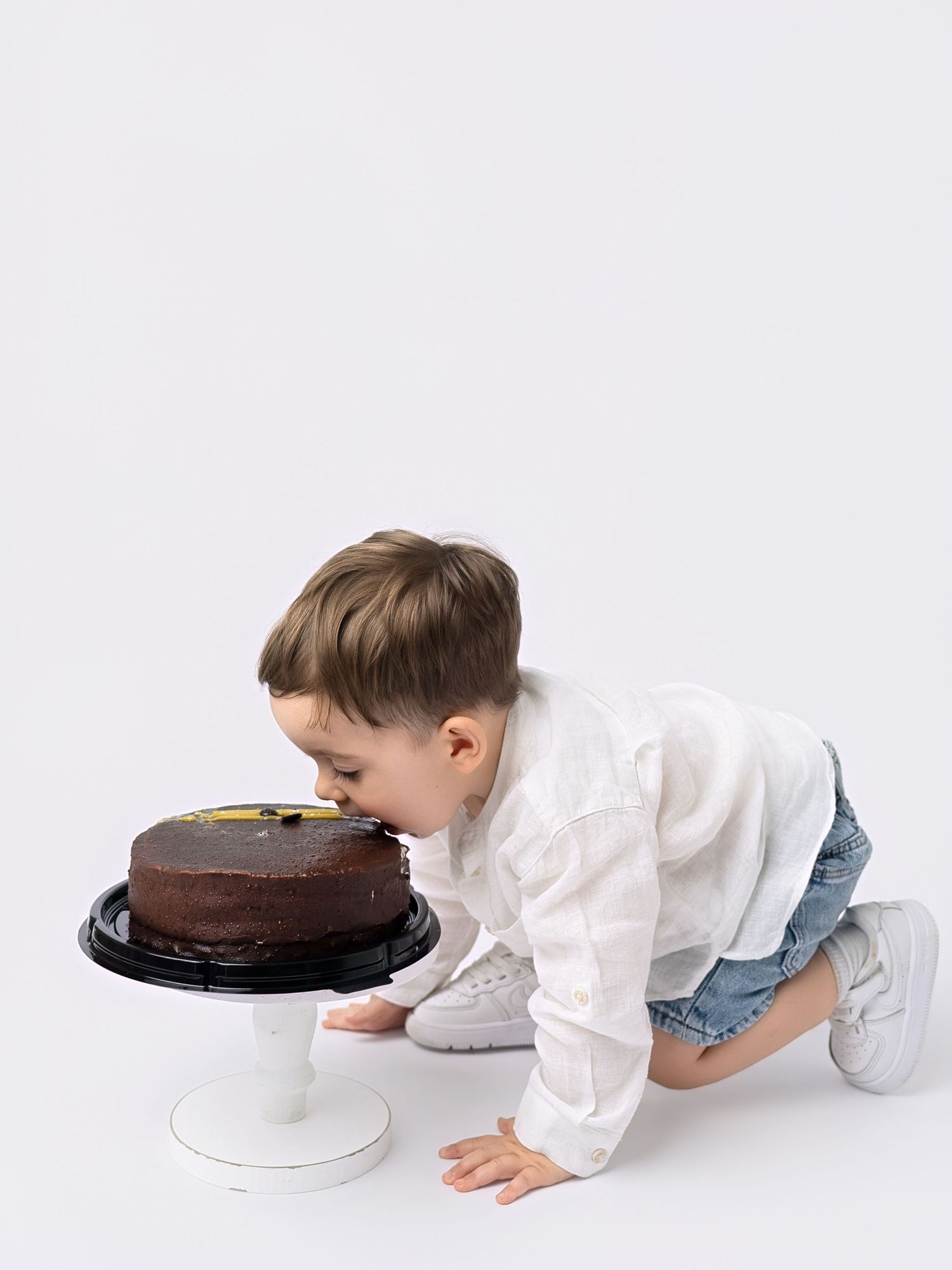 Young boy in white shirt leaning forward to eat chocolate birthday cake on white cake stand.