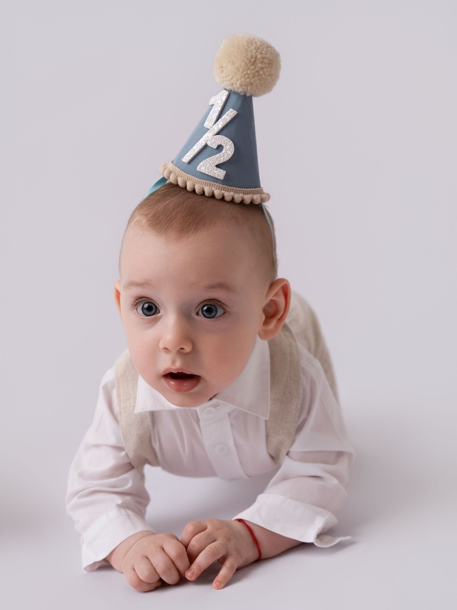 Adorable baby wearing a blue half birthday party hat with pom-pom, lying on tummy in white shirt.