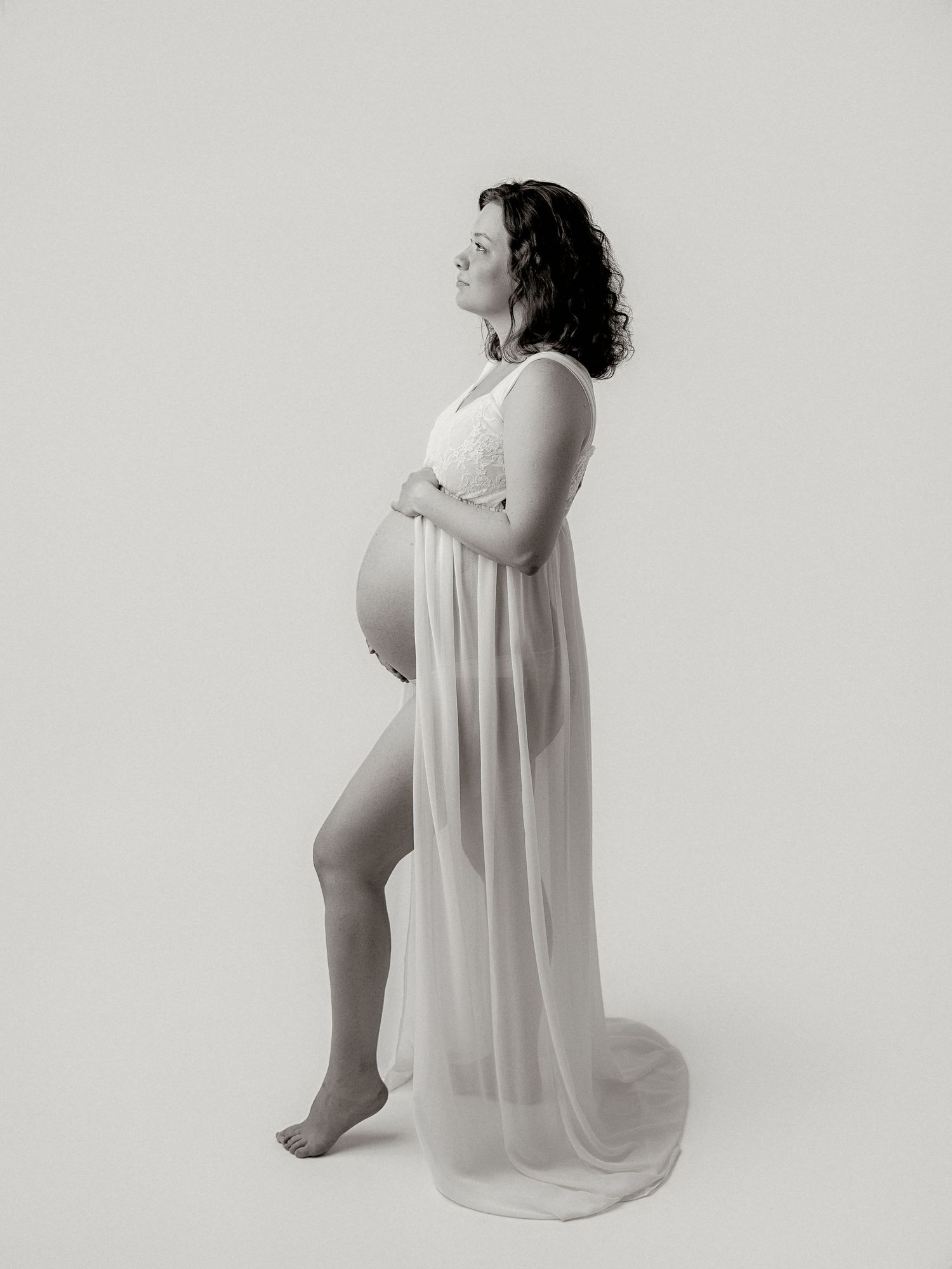 Black and white maternity photo of a pregnant woman in a flowing white gown, cradling her baby bump in a studio.