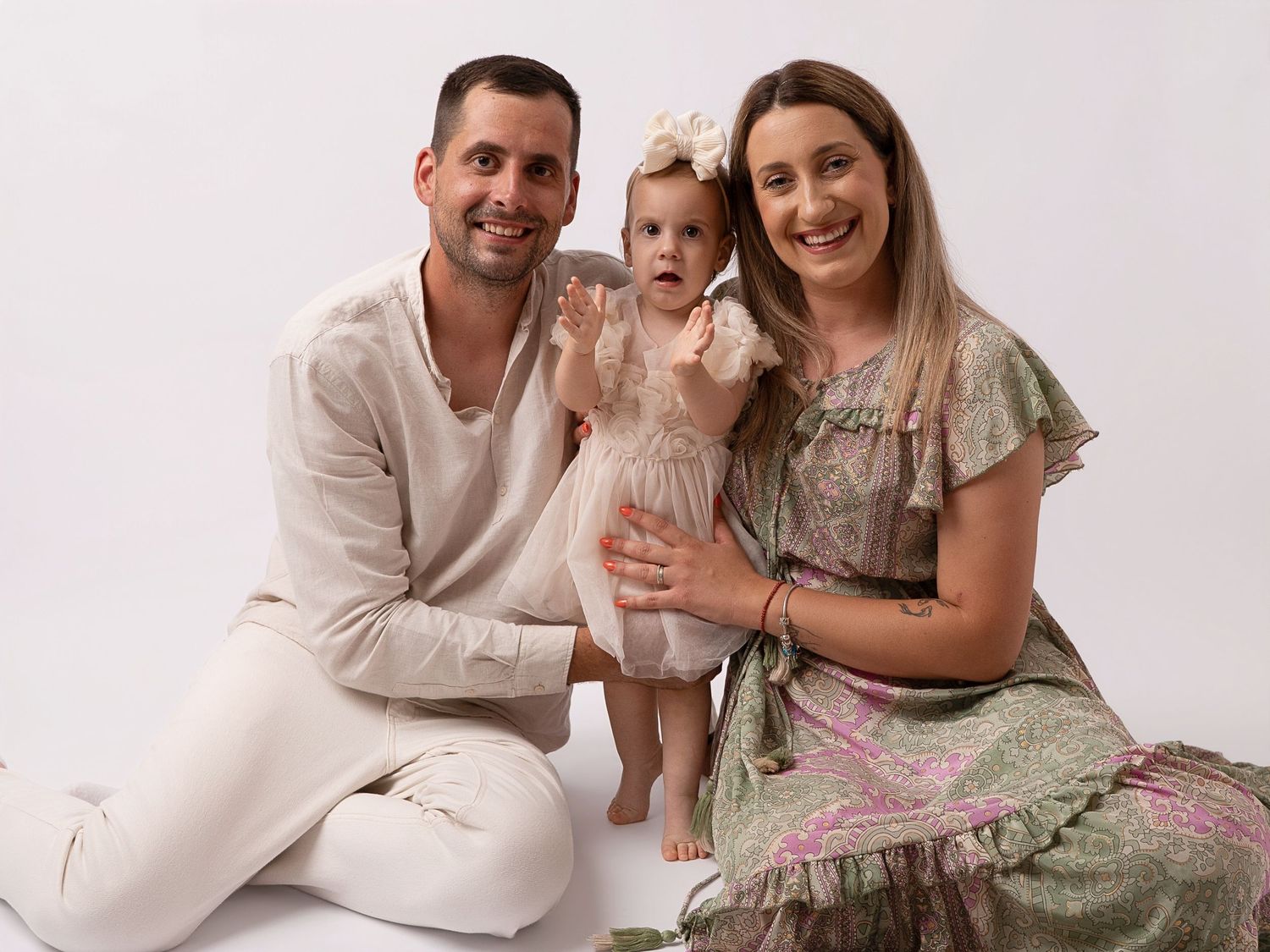 A smiling family with a man, woman, and baby girl in soft pastel outfits posing together against a white background.