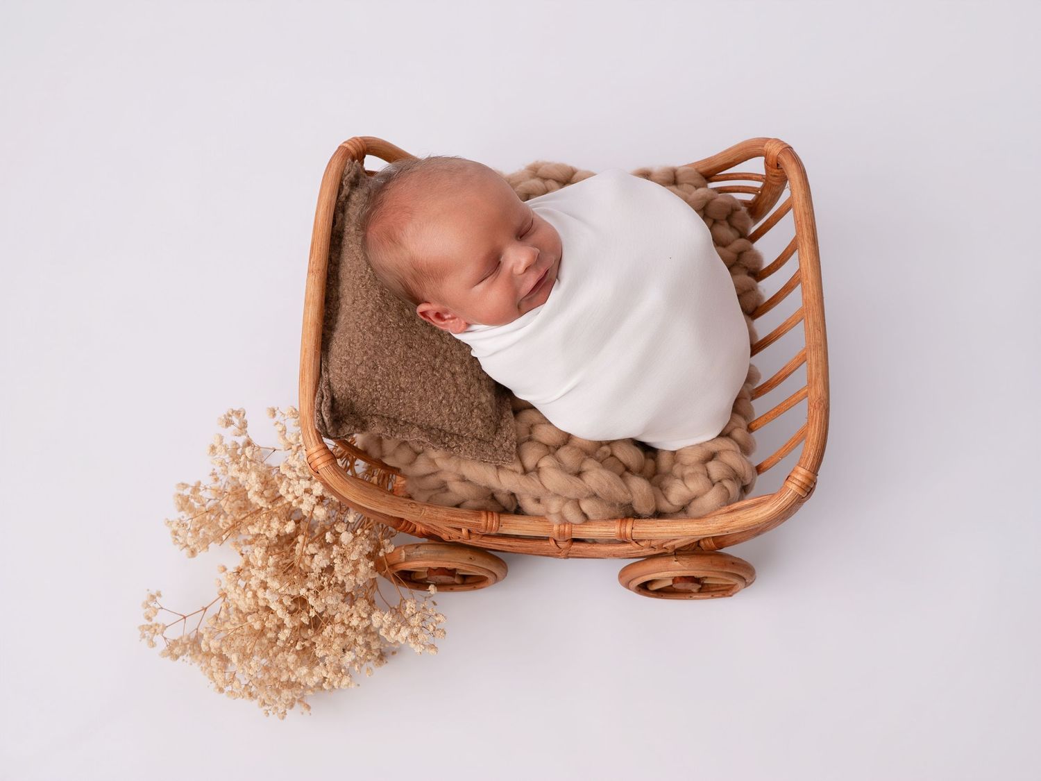 Newborn baby swaddled in white wrap sleeping in a small rattan wagon with dried flowers on white background.
