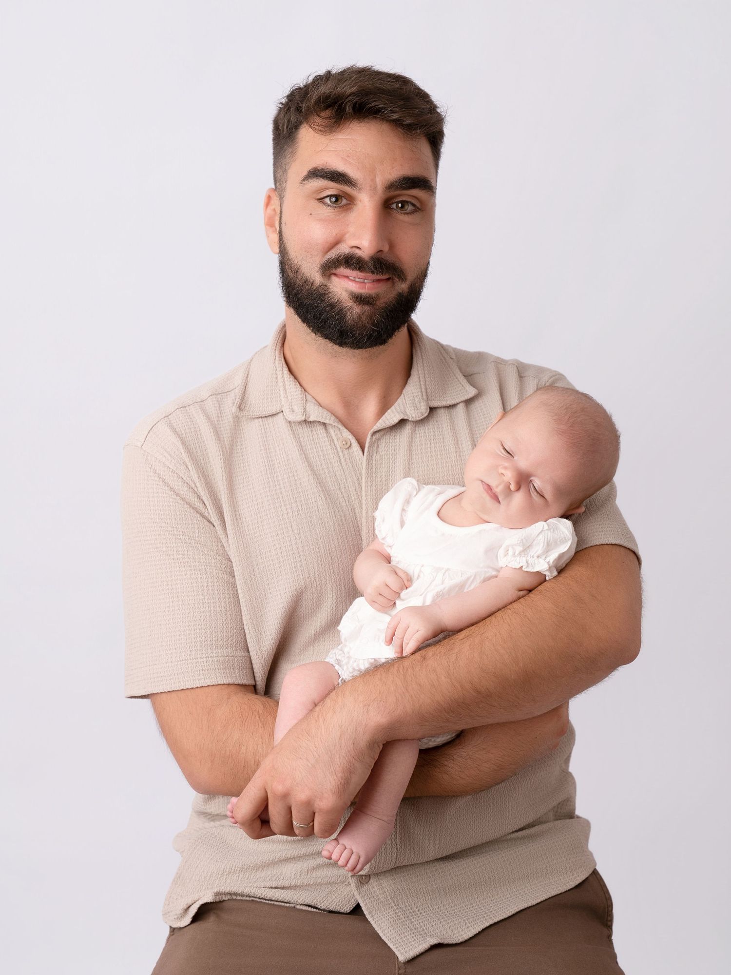 Smiling bearded man in beige polo shirt holding a sleeping newborn baby in white outfit against white background.