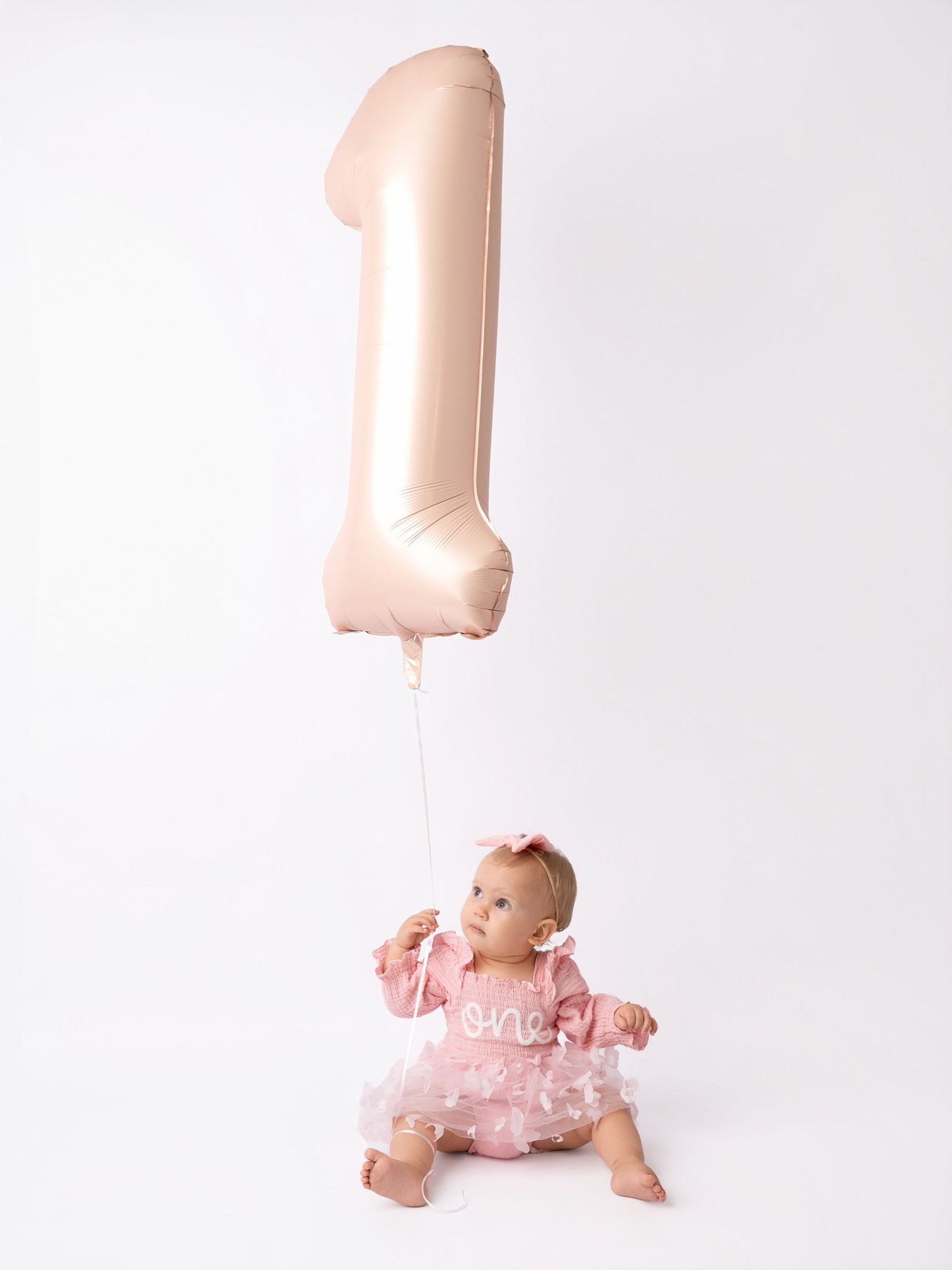 Baby girl in pink tutu dress holds a rose gold number 1 balloon for her first birthday photo.