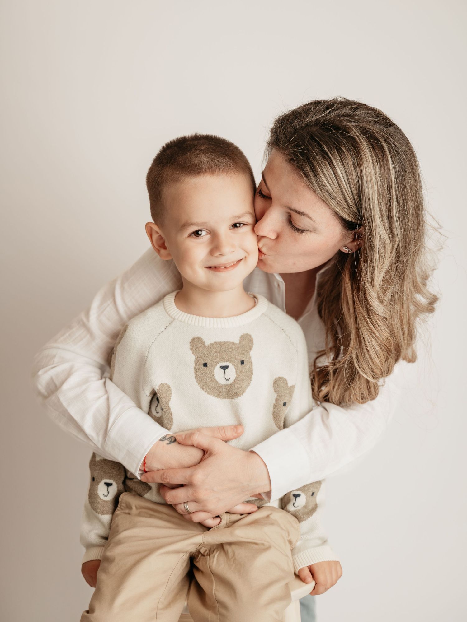 A mother kisses her young son on the cheek as he smiles, both wearing bear-print outfits in a bright studio.