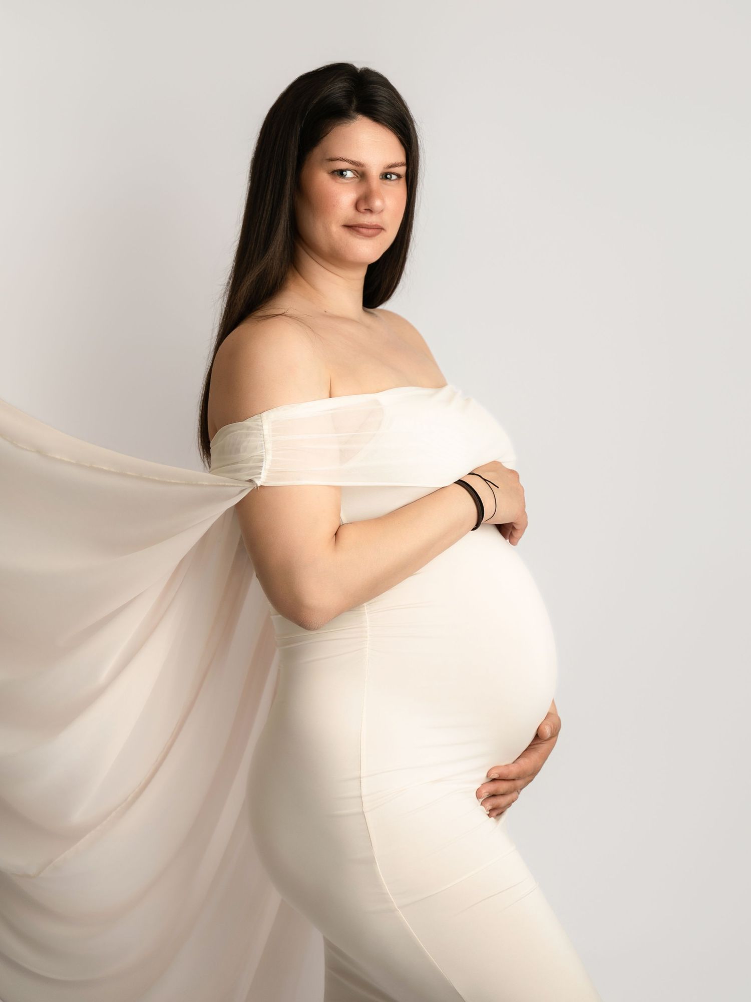 Pregnant woman in elegant off-shoulder cream dress cradling baby bump in professional maternity studio photo.