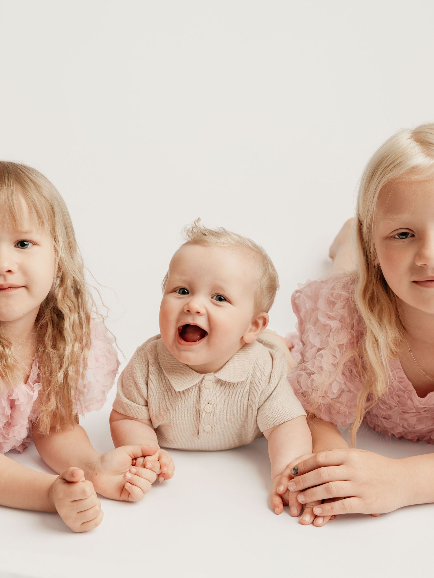 Three young siblings lying together with happy baby in middle in bright white studio portrait.