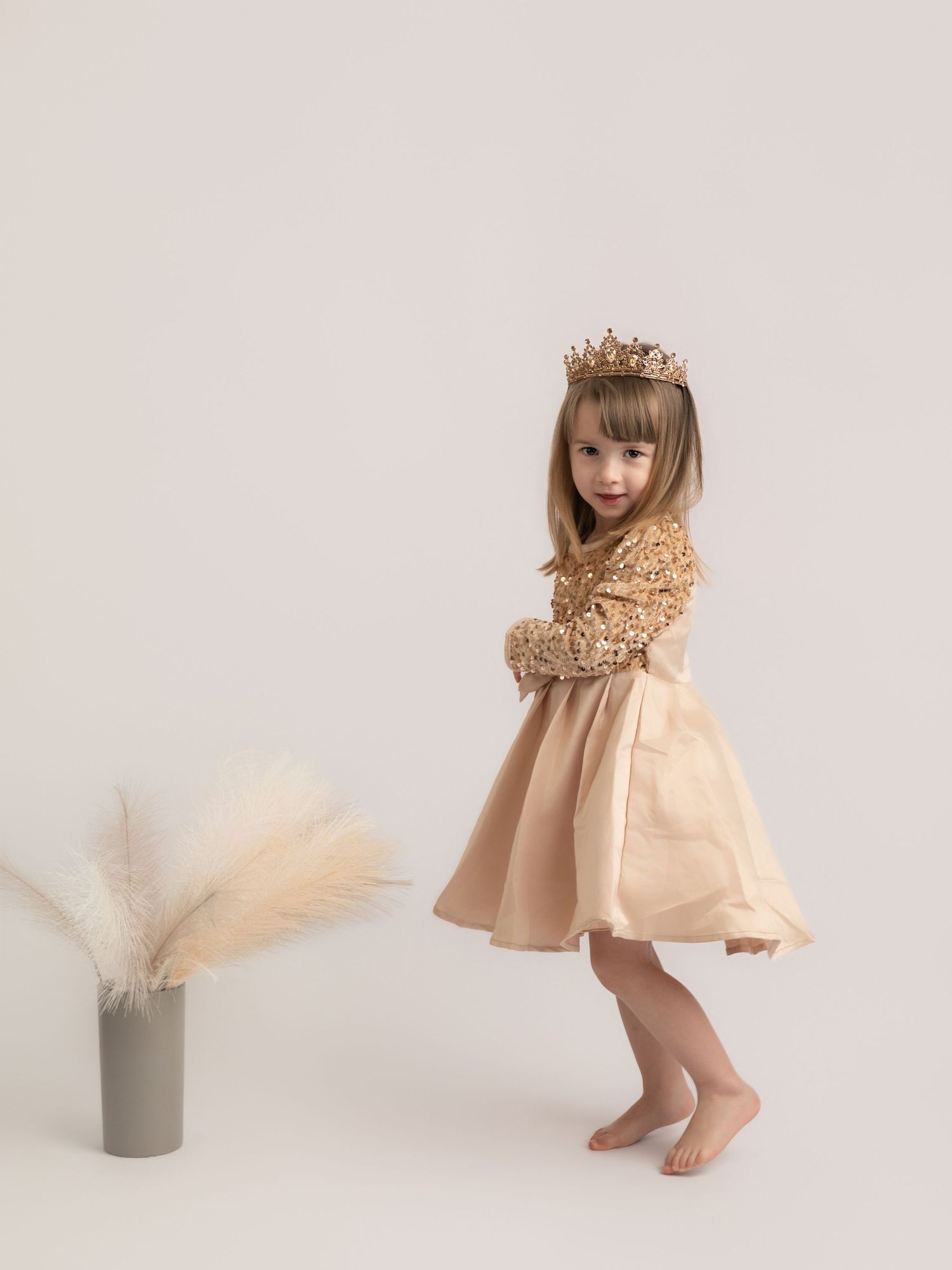 A young girl twirls in a gold sequin and champagne satin dress wearing a crown next to pampas grass decor.