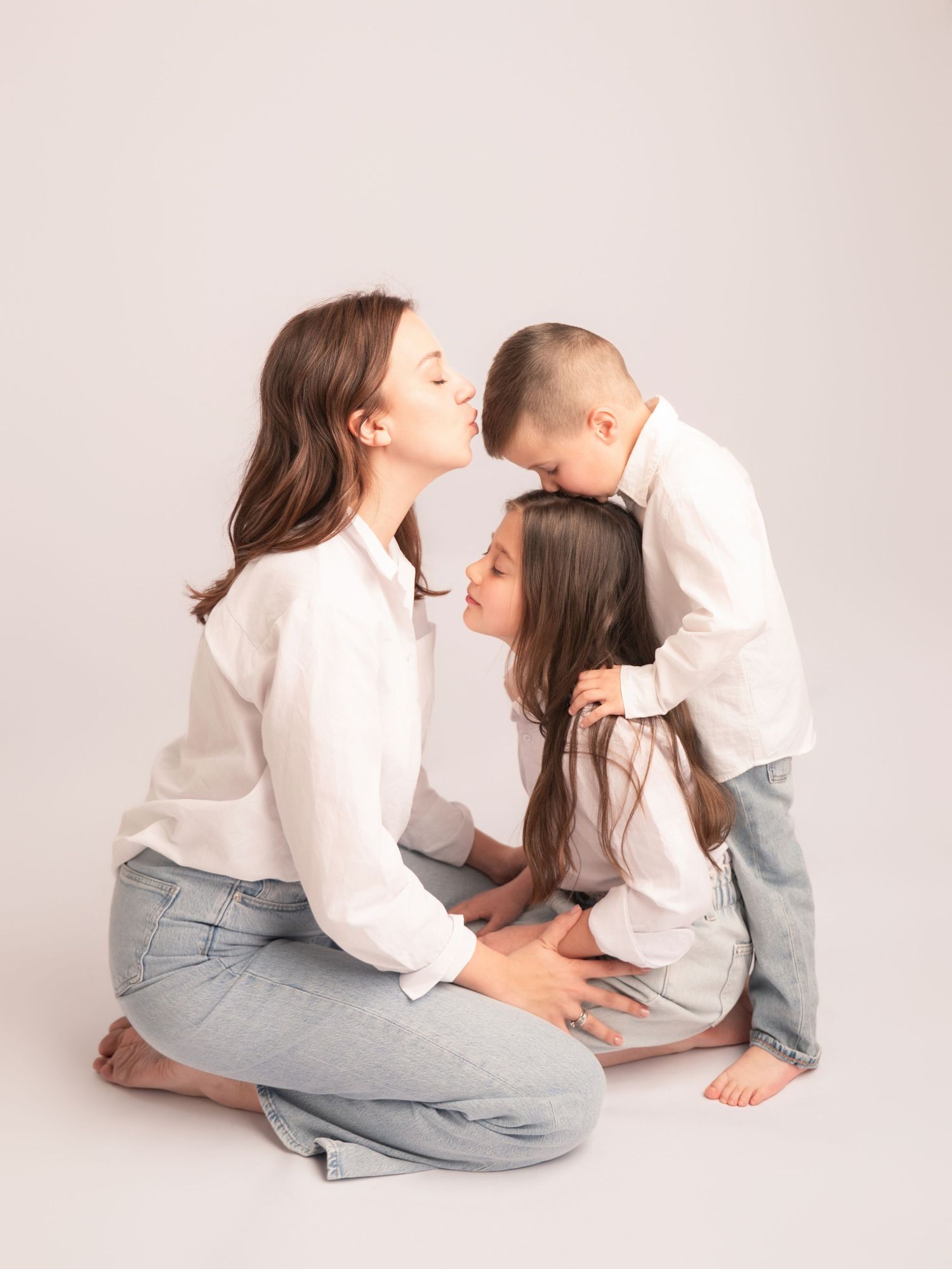A mother shares a loving moment with her two children in white shirts and jeans in a studio photo.