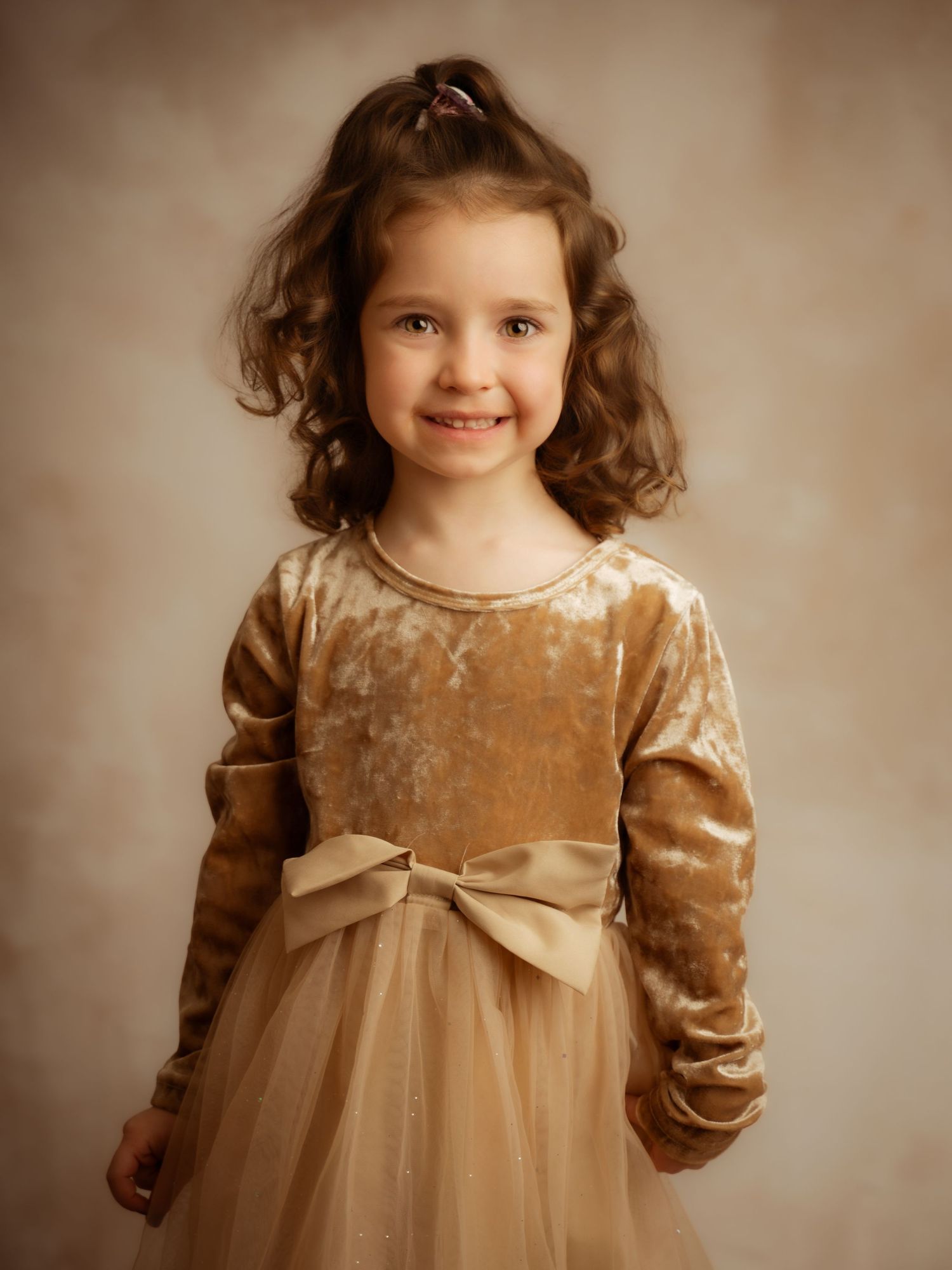 Smiling young girl with curly hair wearing a golden velvet and tulle dress with bow.