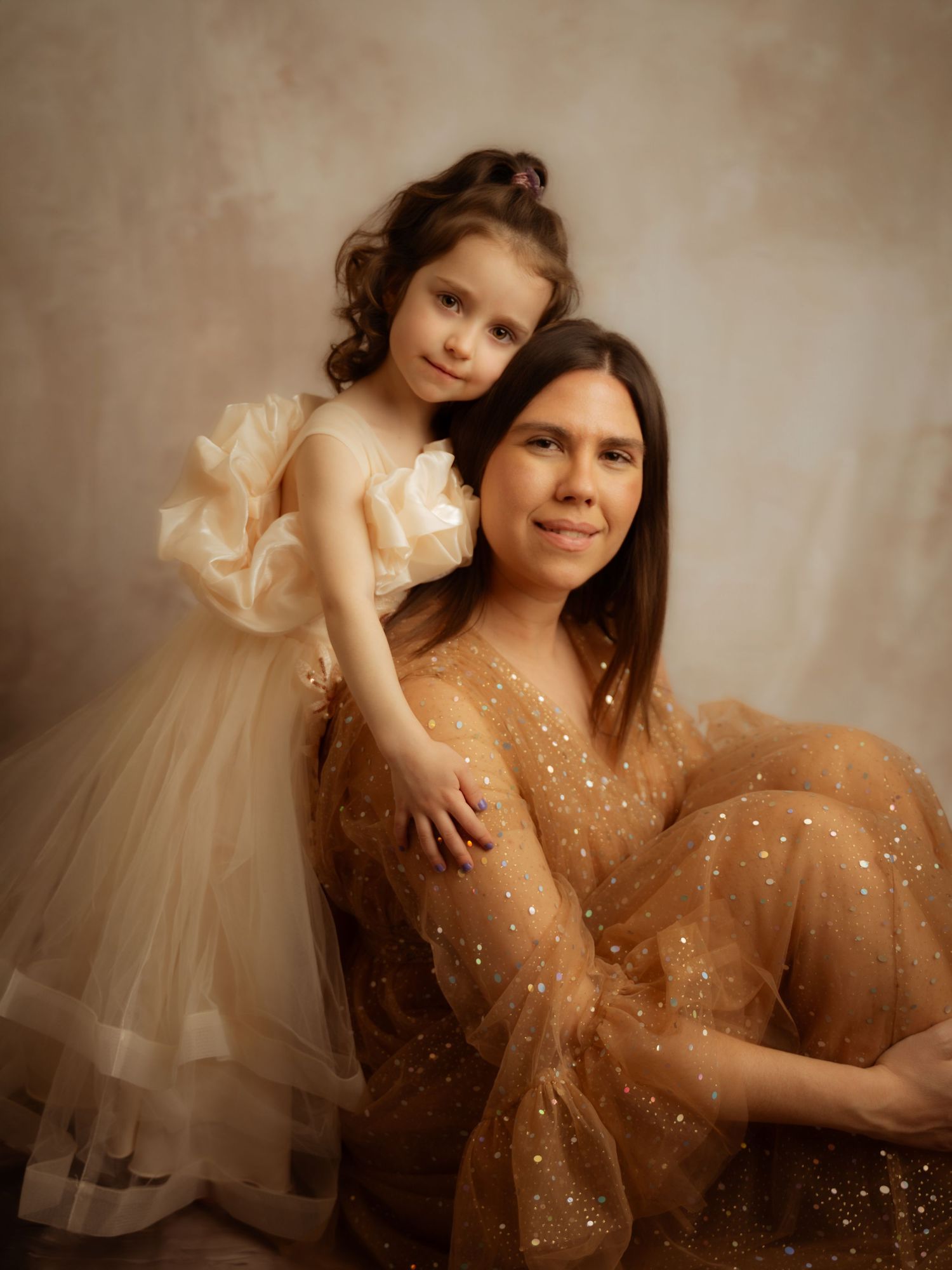 Young girl in cream tulle dress embracing seated woman in gold sparkle gown in studio.