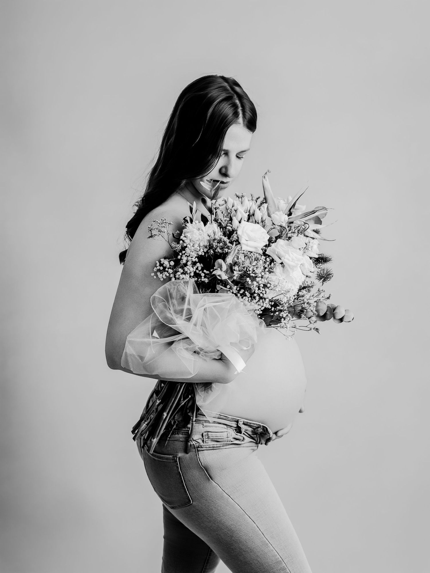 Black and white maternity photo of pregnant woman holding floral bouquet, wearing jeans, looking down tenderly.