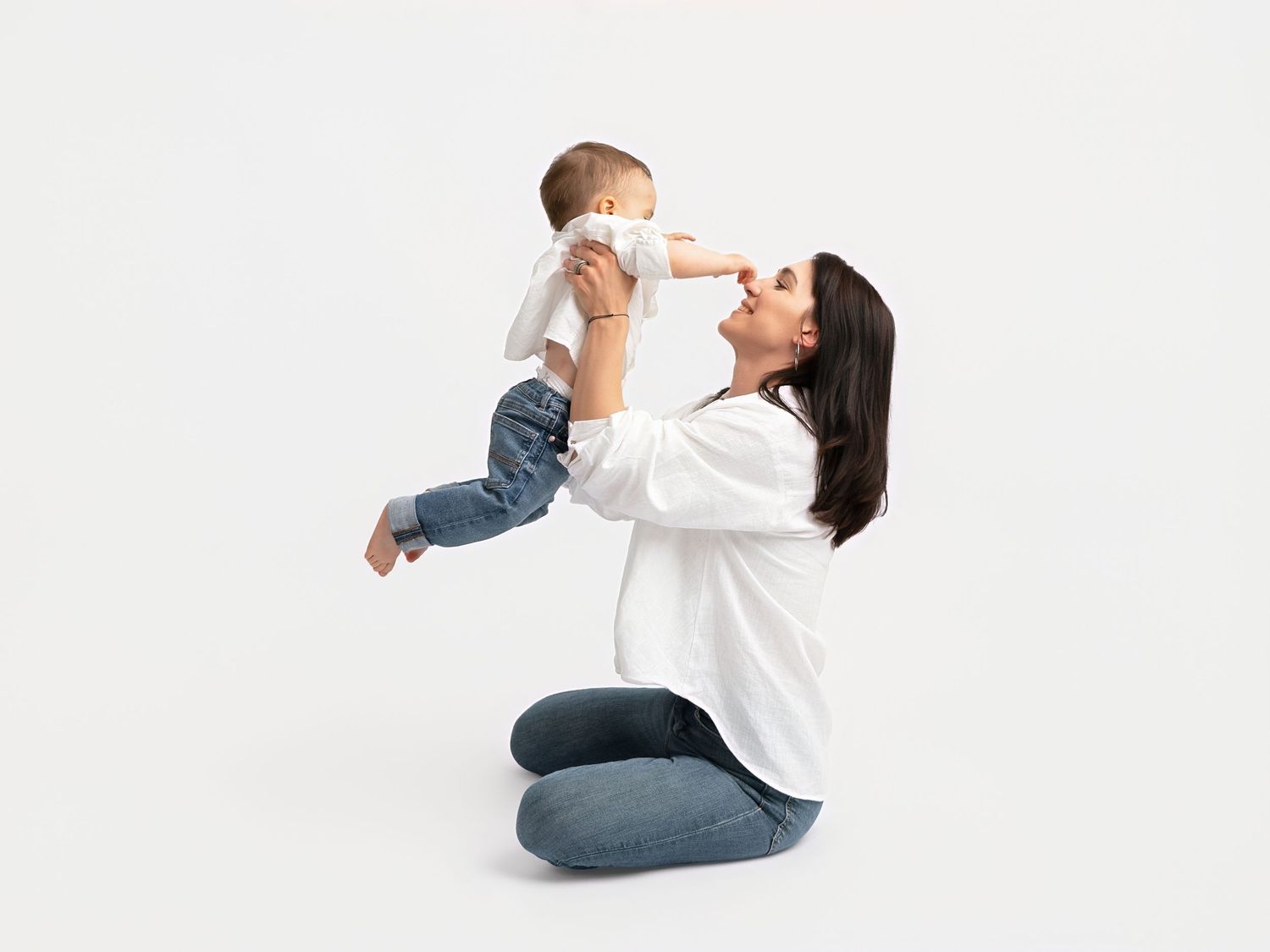 A happy mother kneeling on the floor, lifting her smiling baby in the air against a white background.
