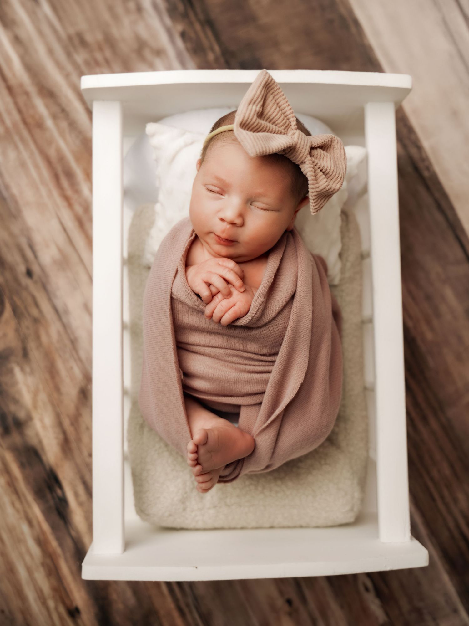 Sleeping newborn baby wrapped in dusty rose swaddle with tan bow headband in white mini crib.