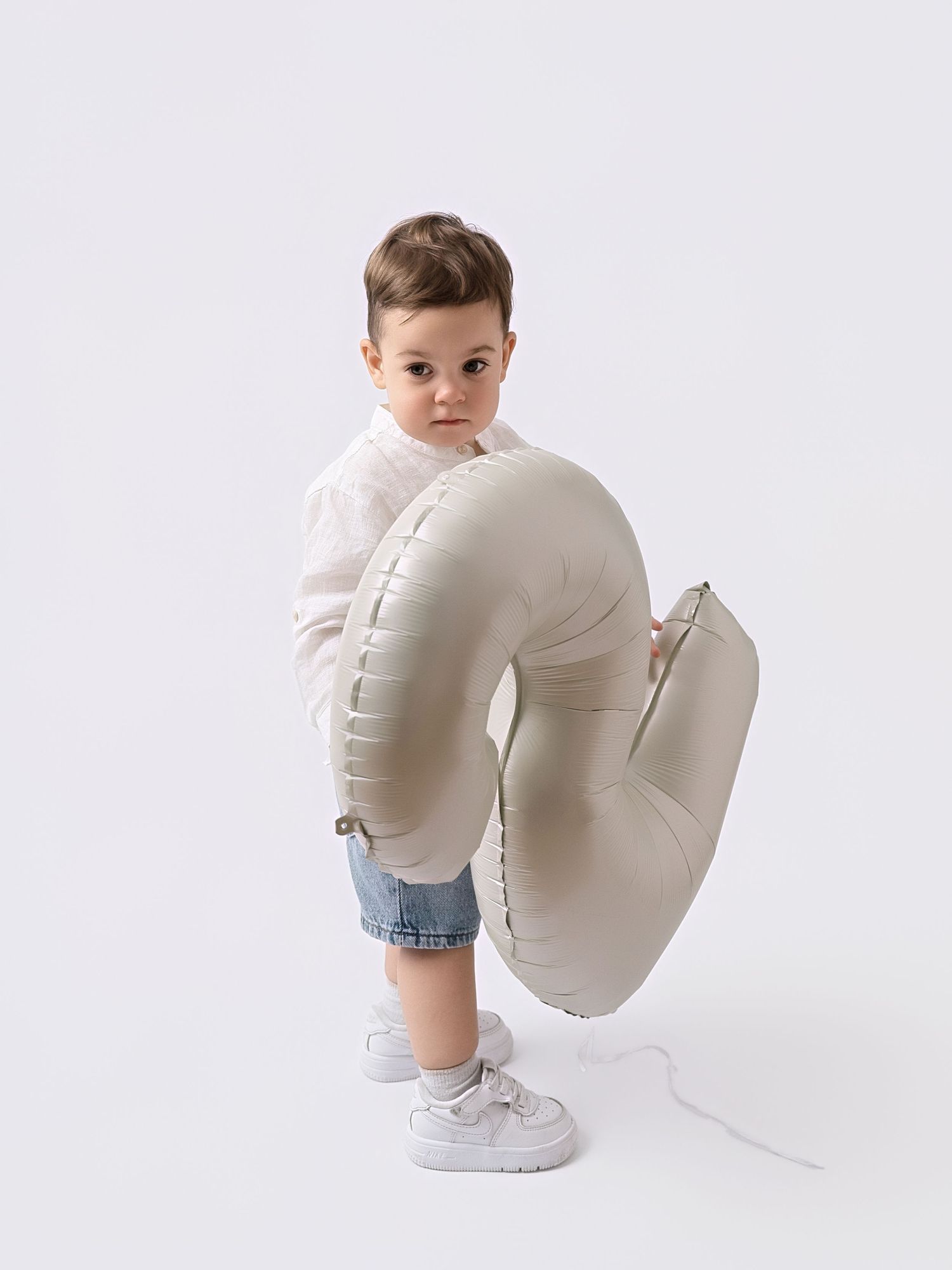 Toddler boy in white shirt hugging a large number 2 balloon on white background.